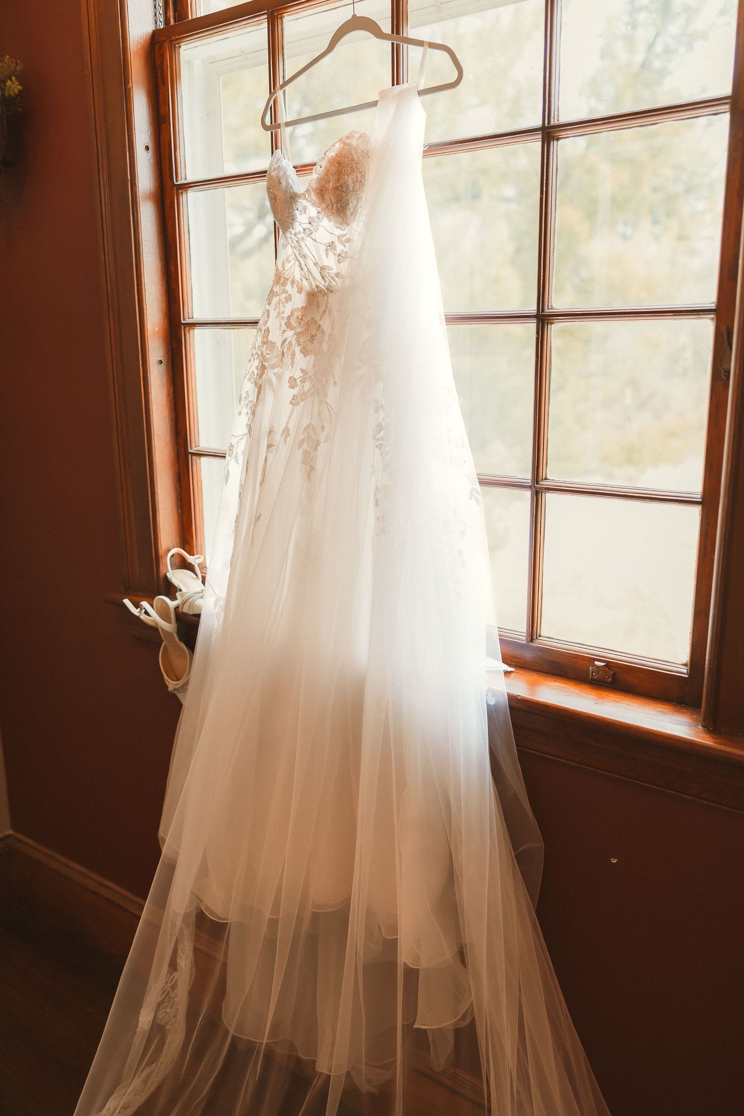 An elegant white lace wedding dress hangs in front of a window with natural lighting.