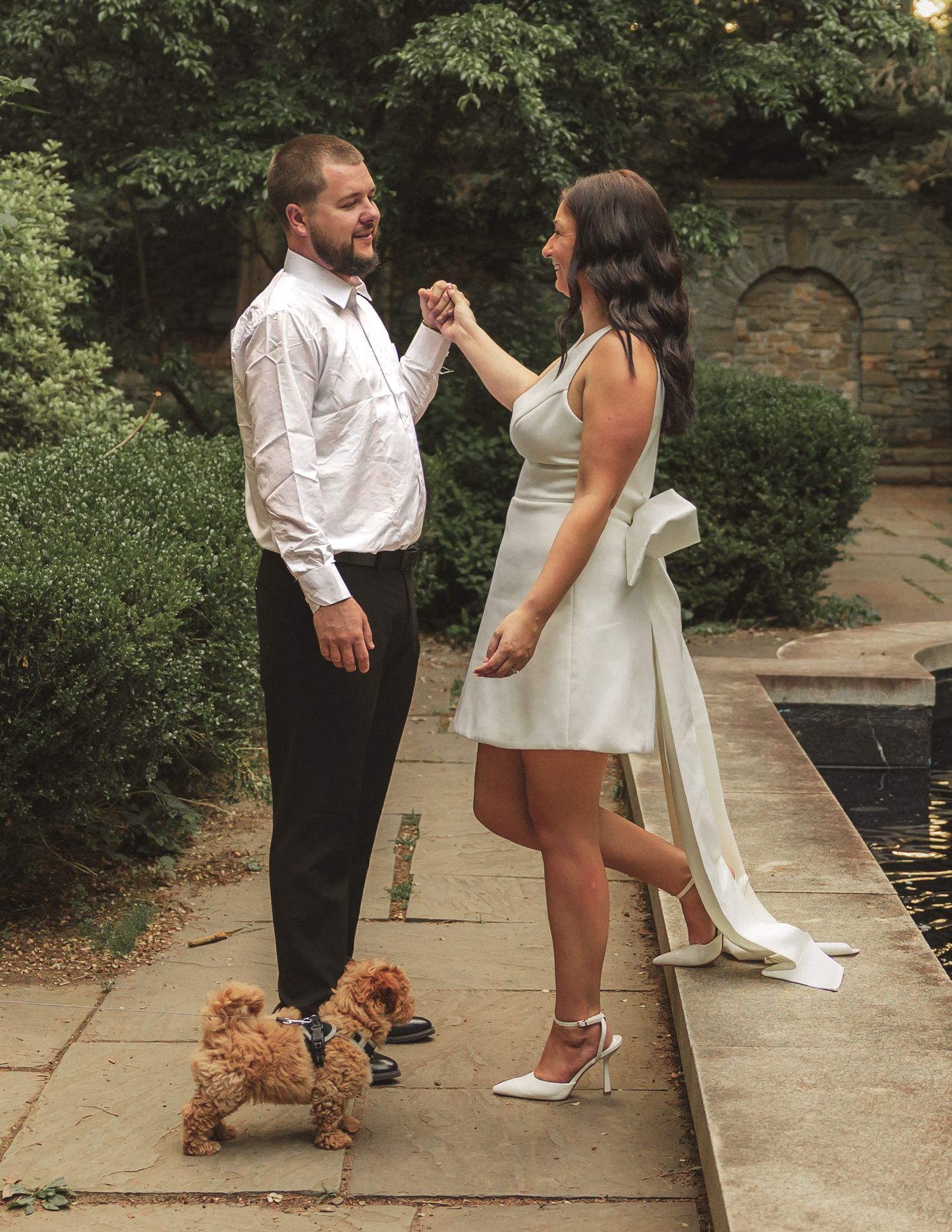 A couple in formal attire shares a moment in a garden setting with a small brown dog on a leash.