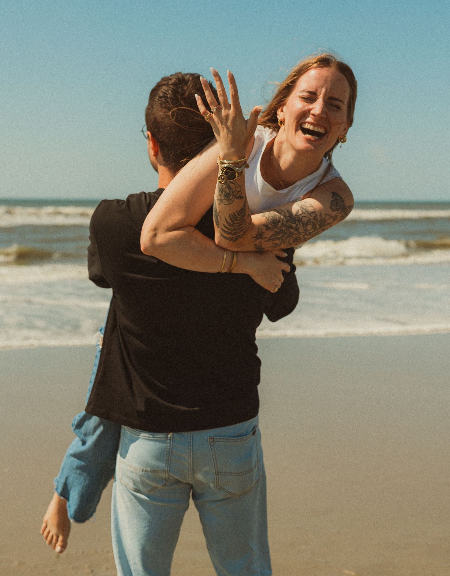 Two people share a joyful moment playing and laughing together on a sunny beach with ocean waves in the background.