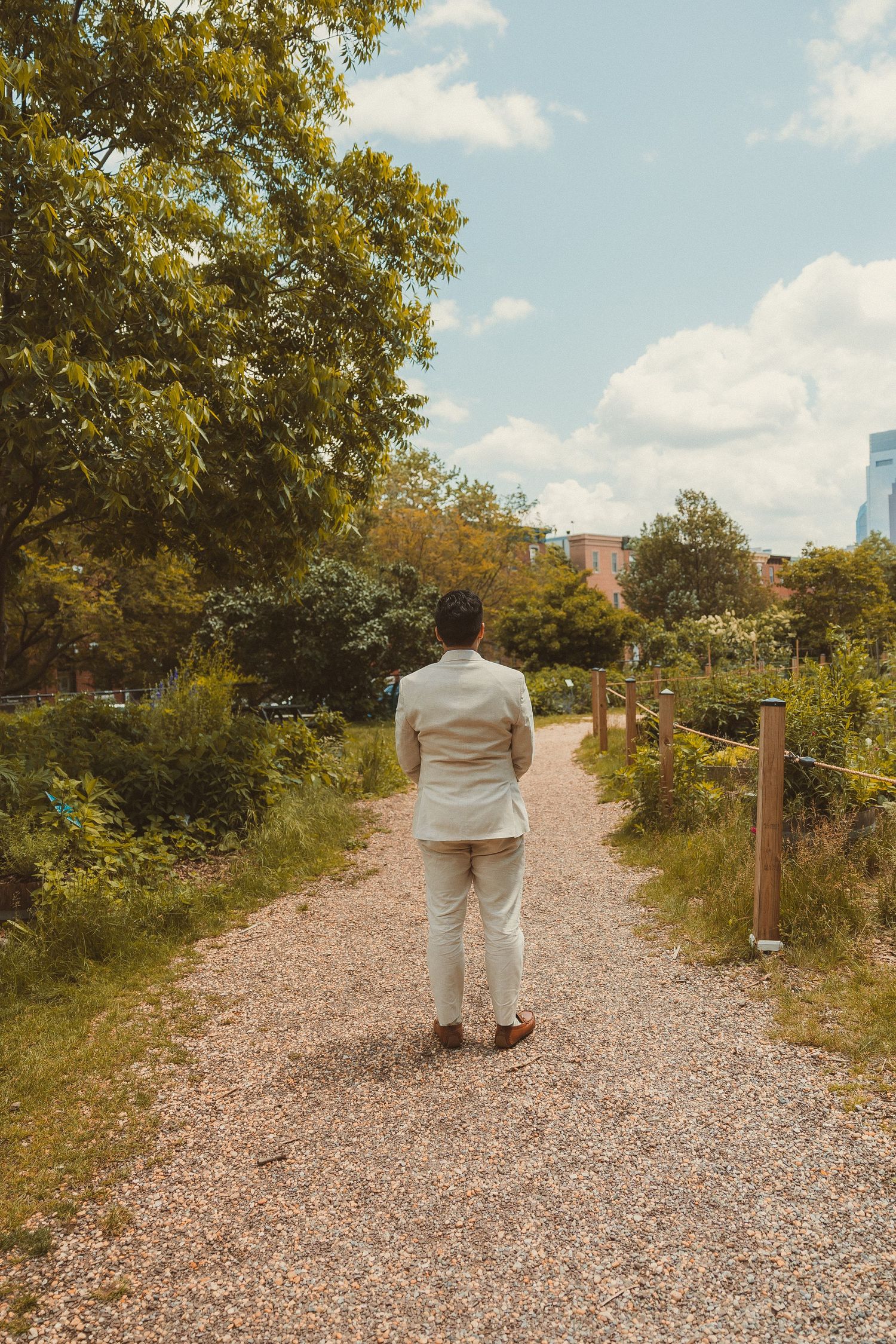 Person in white attire walking alone on a gravel path through a lush garden with city buildings in background.