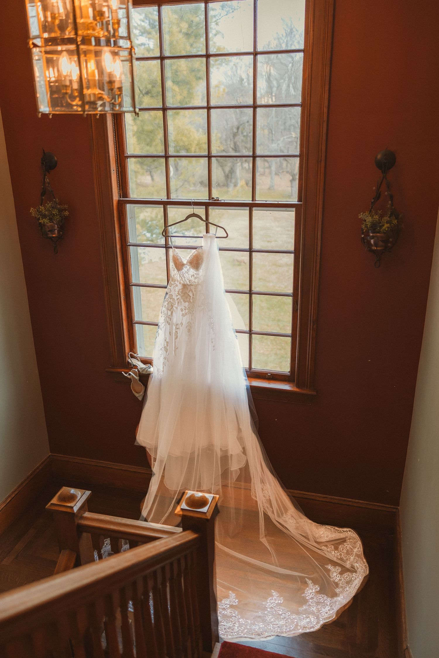 A wedding dress hangs in a window frame with a chandelier and wooden staircase visible.