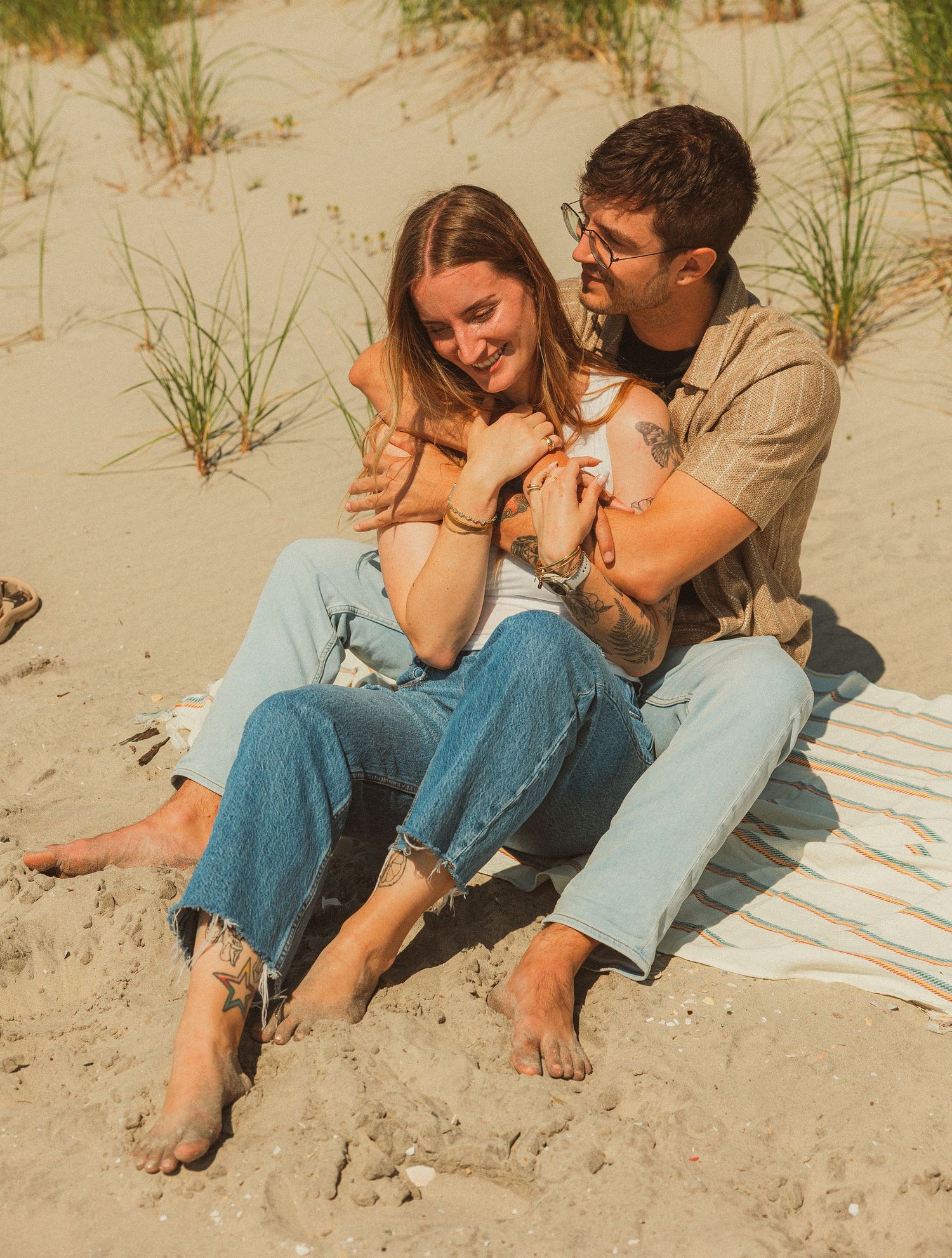 A couple sits together intimately on a sandy beach blanket during a warm sunny day.