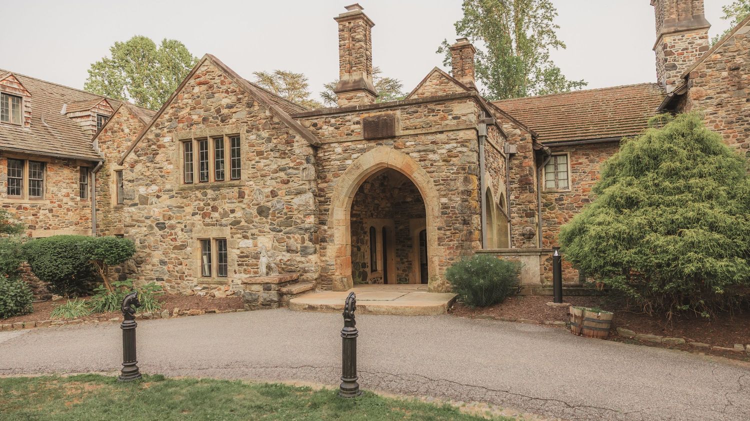 Historic stone archway entrance with Gothic architecture and flanking chimneys at medieval style building surrounded by greenery.
