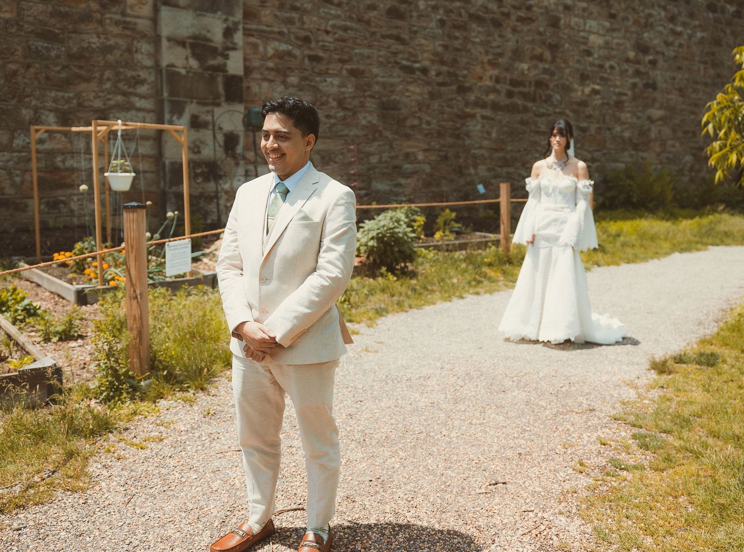 Two people in elegant white wedding attire stand on a gravel path near a rustic brick wall with hanging lanterns.