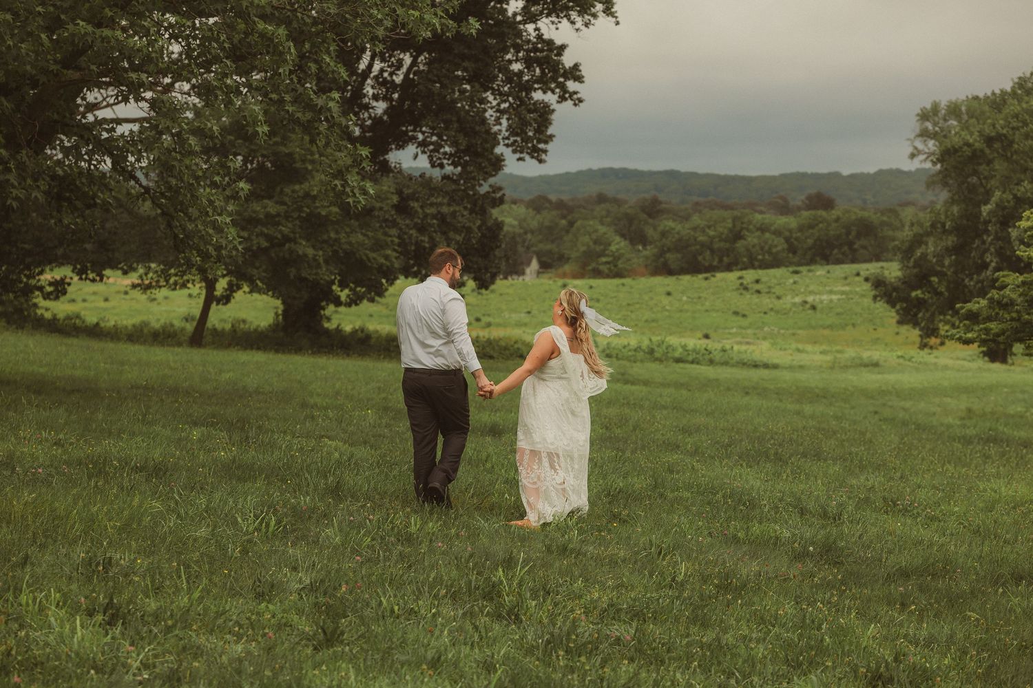 A scenic wedding photo series showing a couple walking together across a lush green hillside meadow on an overcast day.