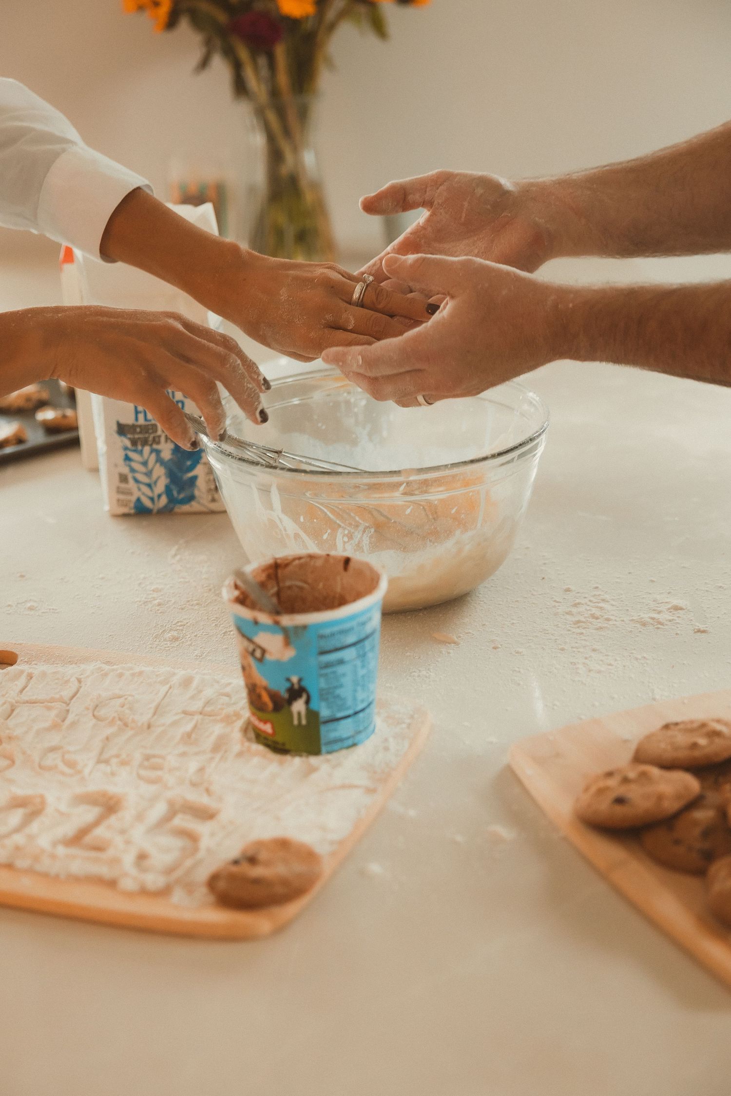 Hands kneading cookie dough together on a flour-dusted white surface with an orange flower arrangement in the background.