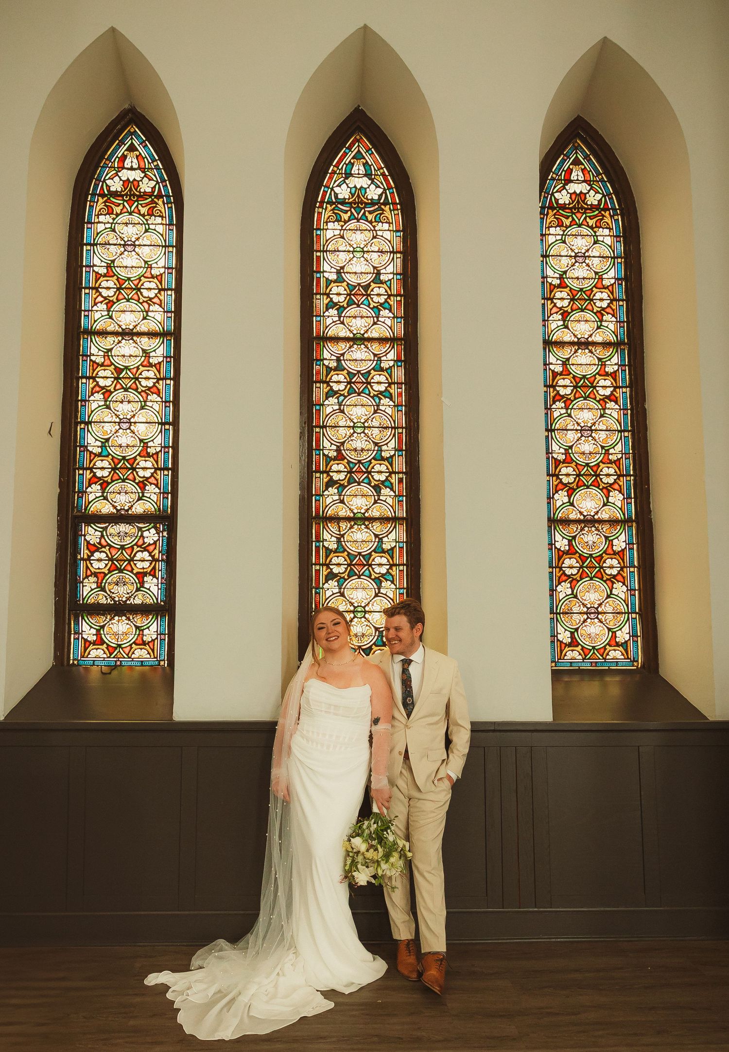A couple stands in front of three tall stained glass church windows with beautiful ornate patterns in warm lighting.