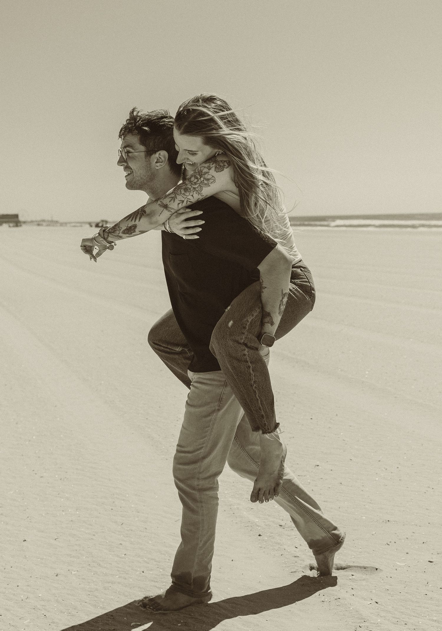 A couple enjoying a carefree moment on a sandy beach with one person giving the other a piggyback ride in sepia tones.