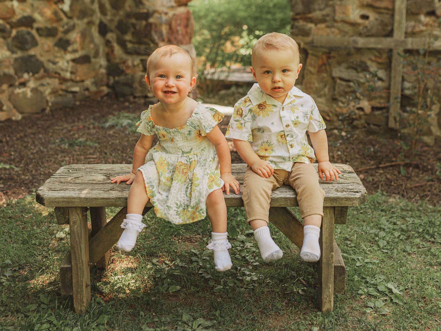Two small toddlers wearing matching floral outfits sit together on a rustic wooden bench in a garden setting.