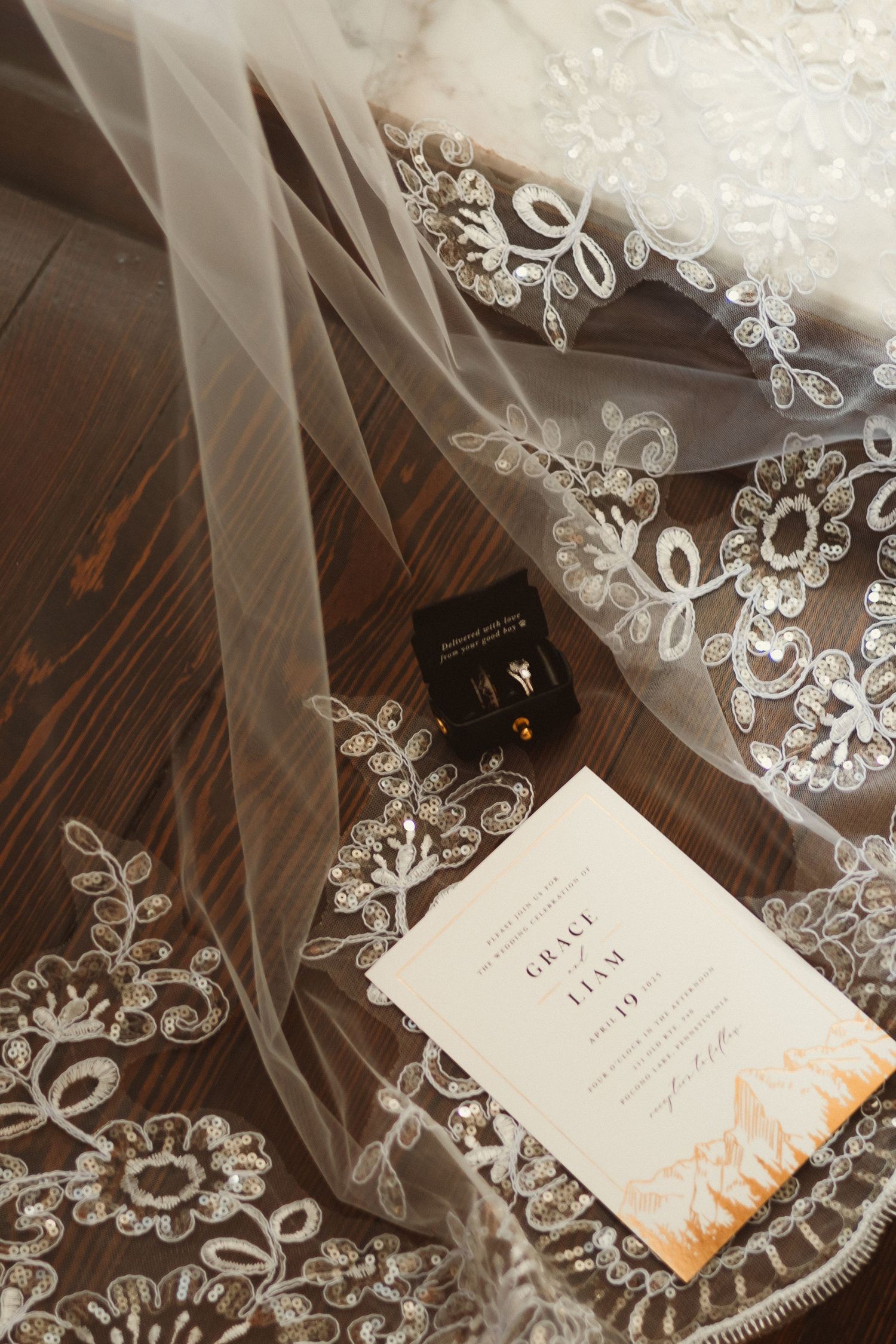 Delicate lace-trimmed wedding veil draped over an invitation card on dark wooden flooring.