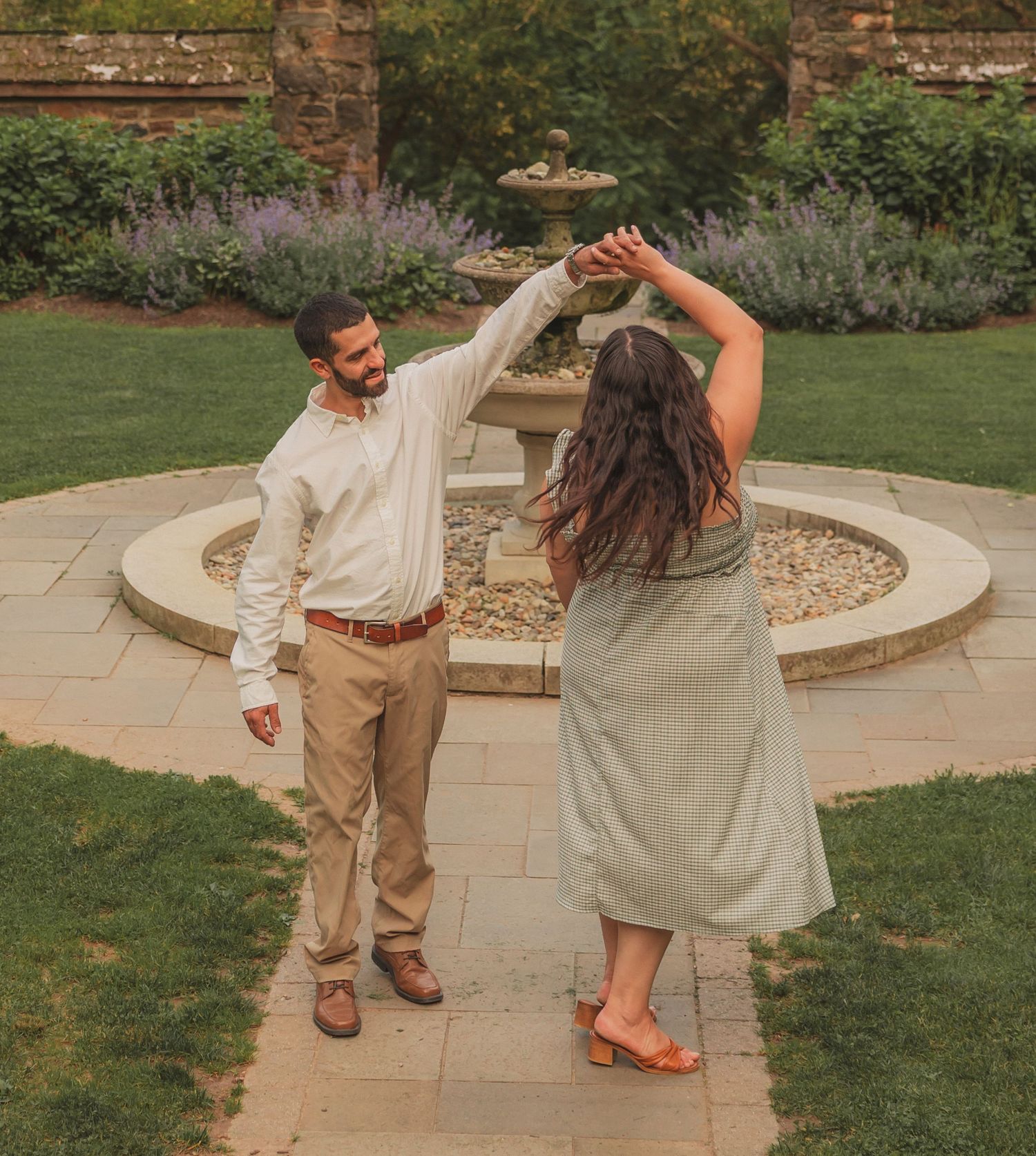 A couple dances together on a stone patio with fountain and lavender garden in the background during golden hour.