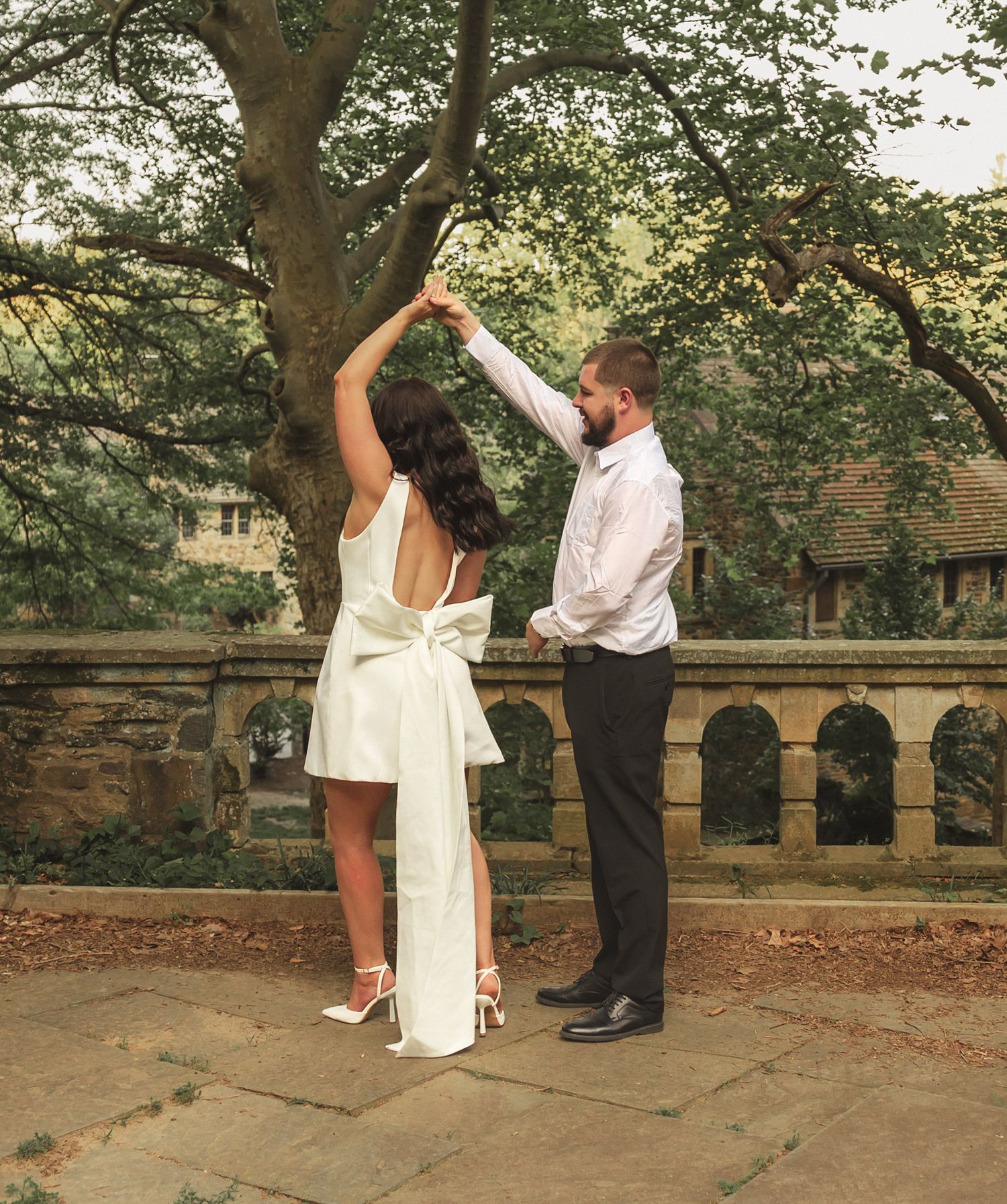 A couple dances together in an outdoor park setting with stone arches and trees in the background.