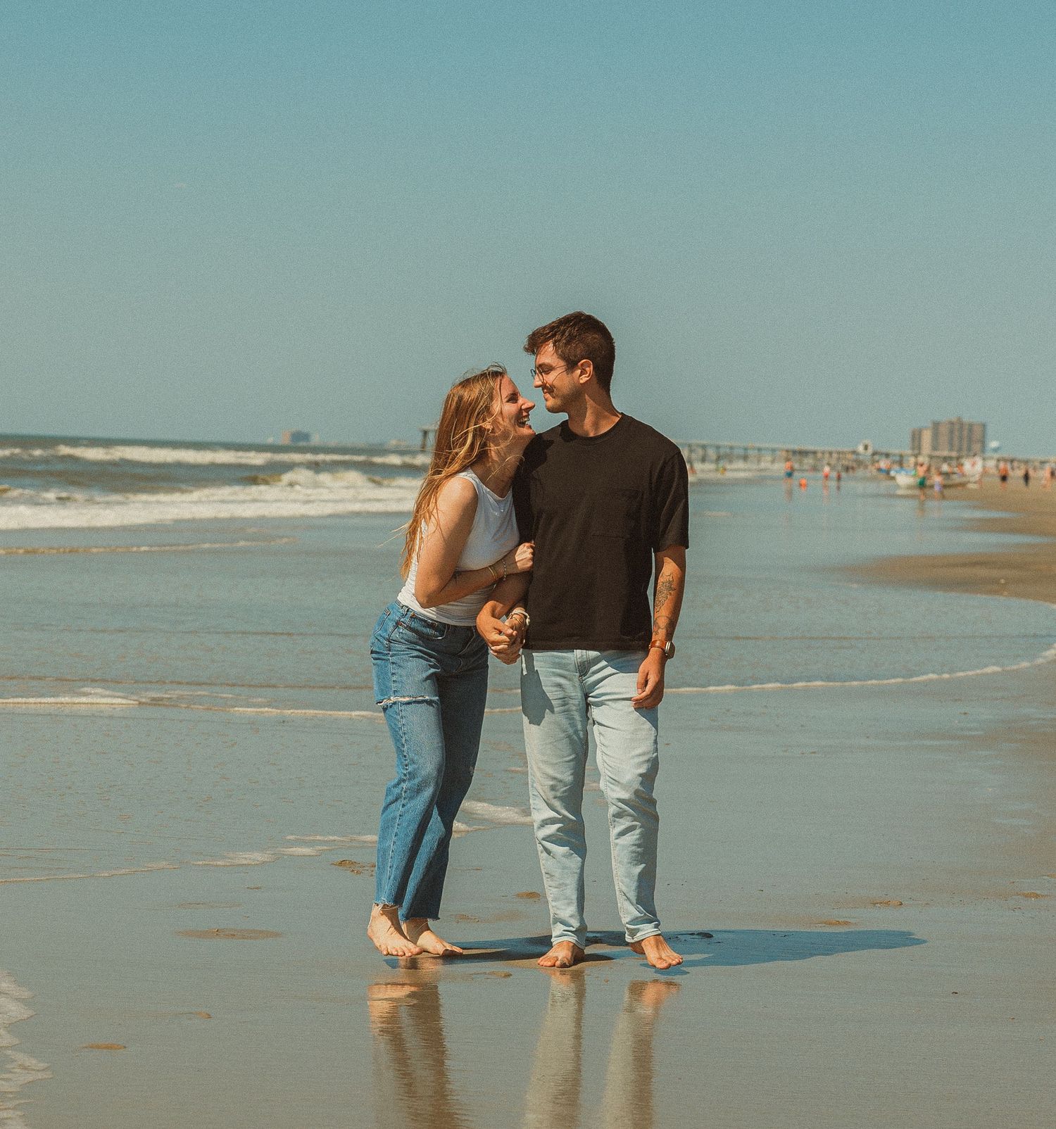 A couple embraces while standing barefoot on a sunny beach with coastal buildings in the background.