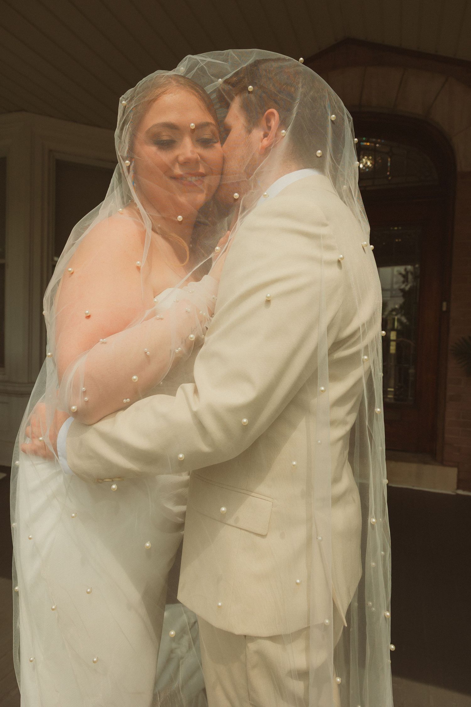 A tender moment between newlyweds as they embrace under a delicate pearl-dotted veil at their wedding ceremony.