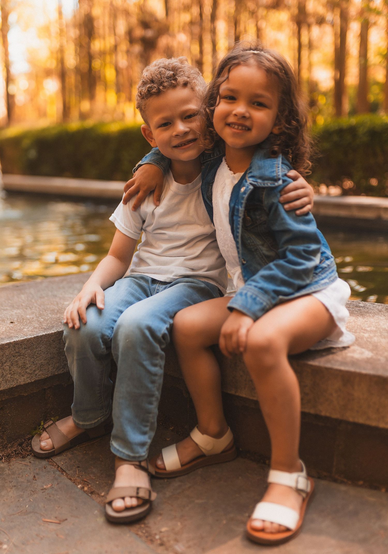Two children sitting together on outdoor stone steps at sunset in a park with autumn trees in the background.