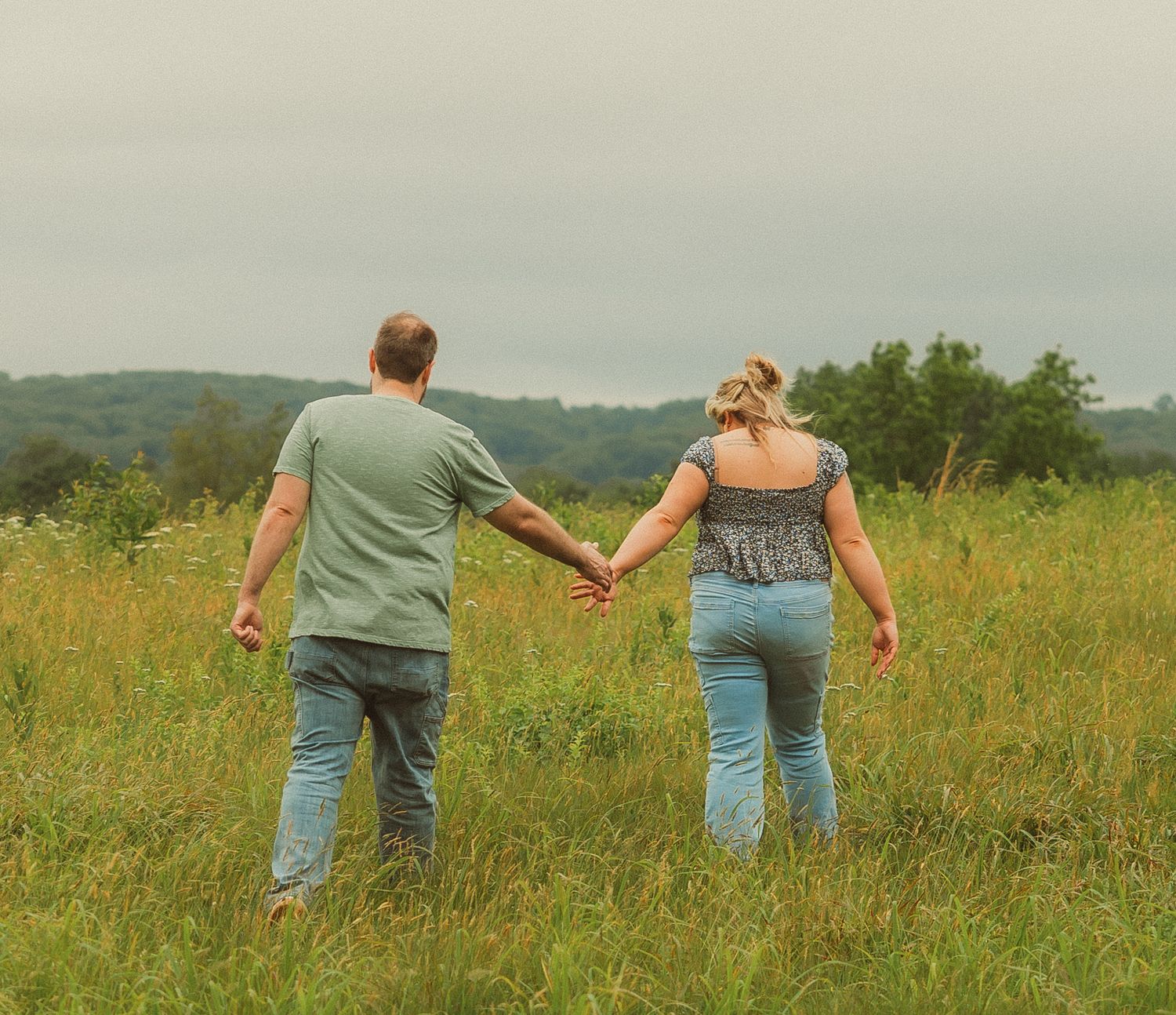 A couple holds hands while walking through a sunny summer meadow with rolling hills in the background.