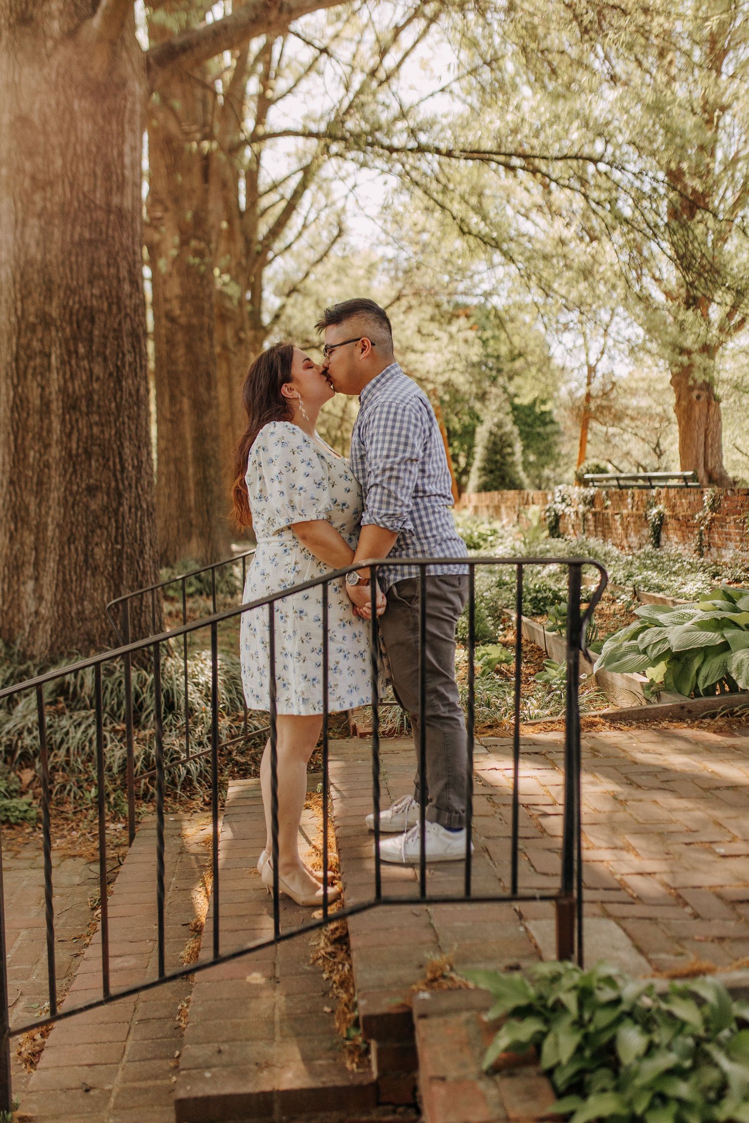 A couple shares a tender moment on wrought iron steps surrounded by tall trees and greenery.