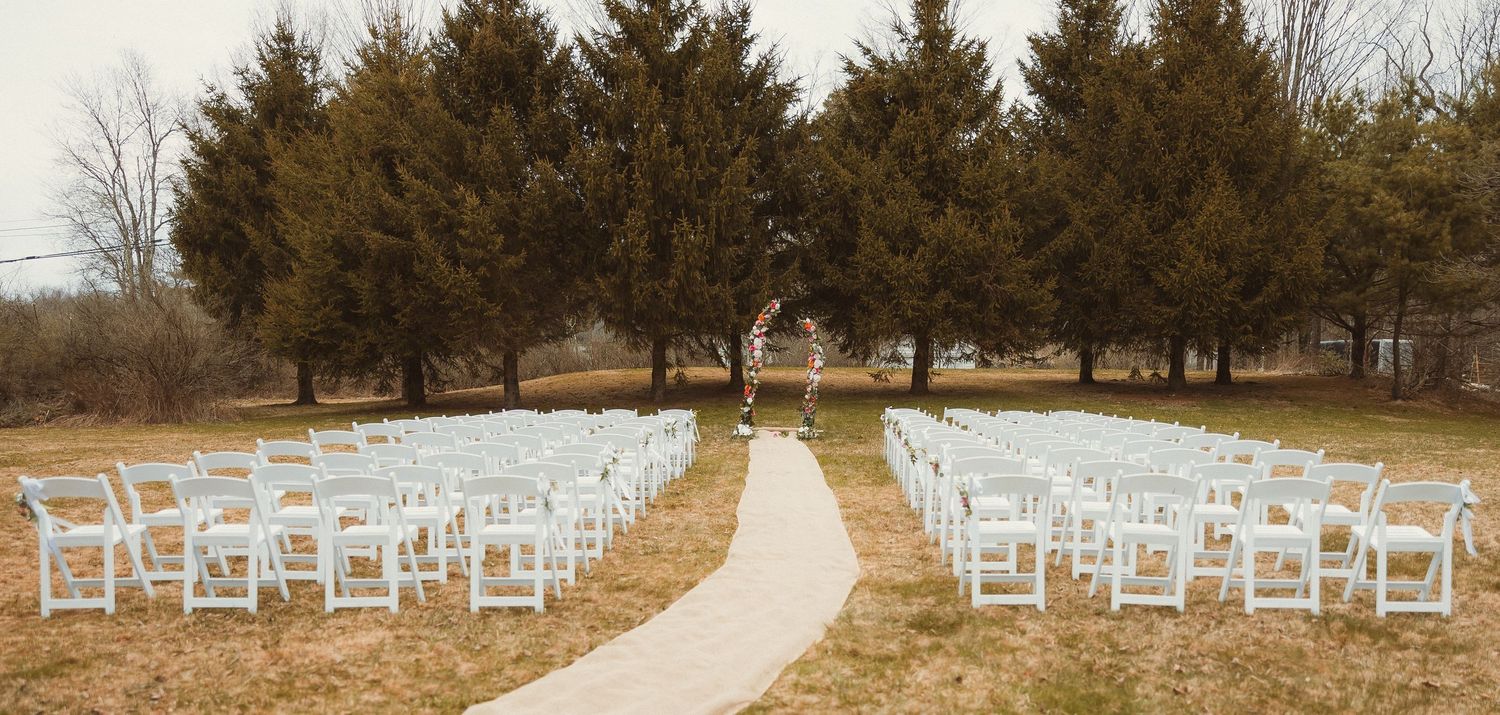 White chairs arranged in rows along a winding path leading to a ceremony space framed by evergreen trees.