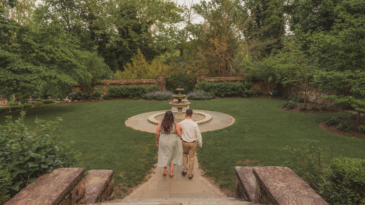 Two people embrace on a curved garden path surrounded by lush greenery and landscaped grounds.