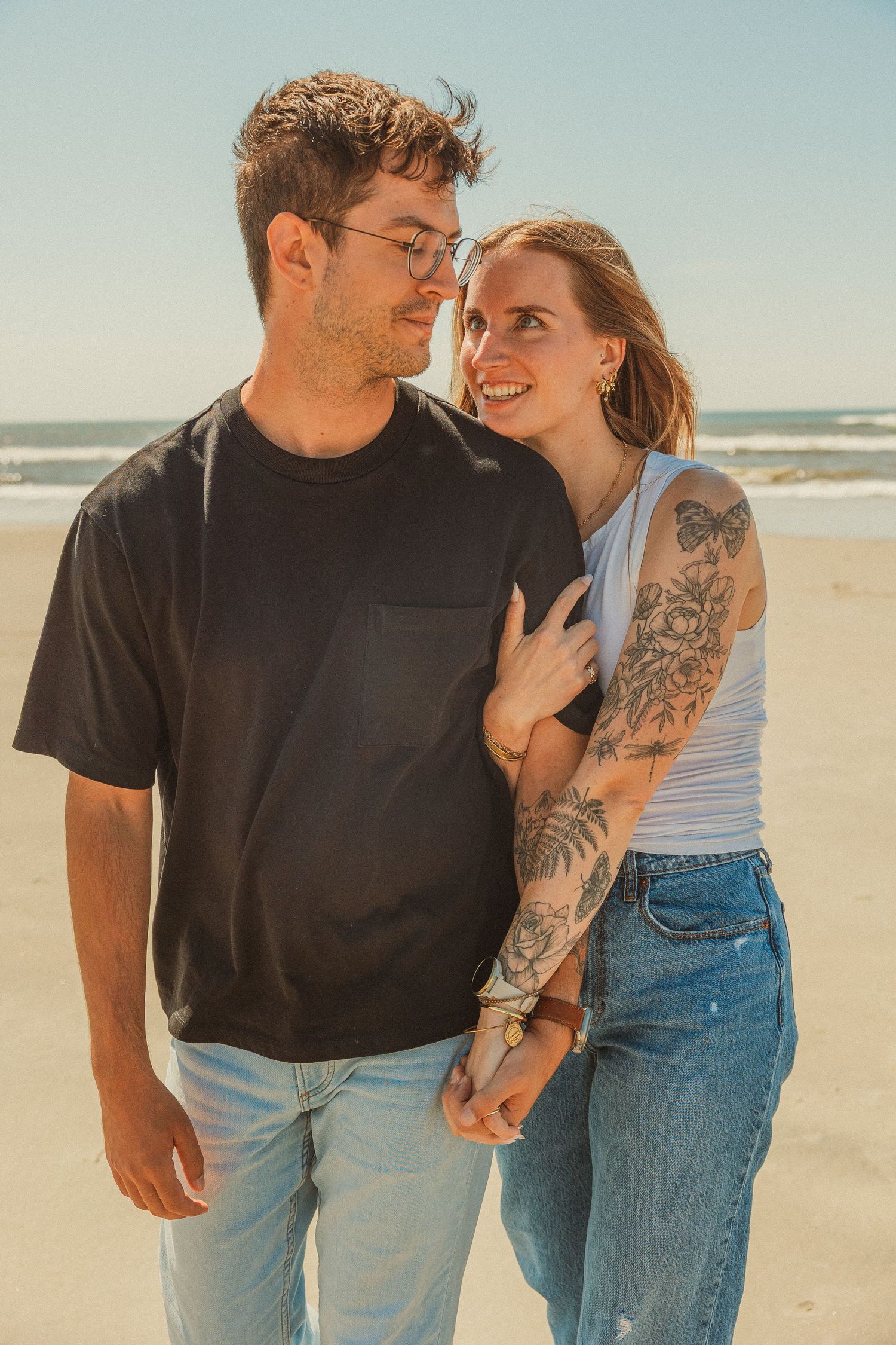Romantic couple embraces on sunny beach, with person wearing glasses and black shirt next to tattooed partner in tank top.
