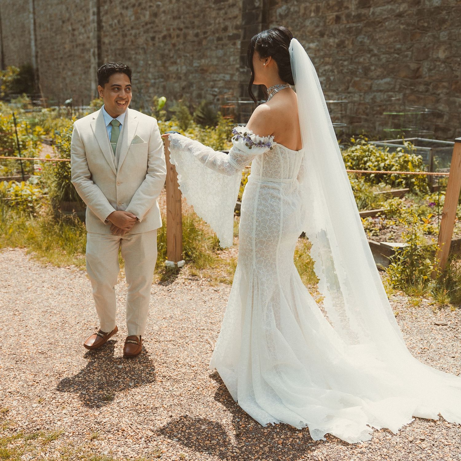 A couple in wedding attire poses outdoors against a brick wall with sunlit vegetation in the background.