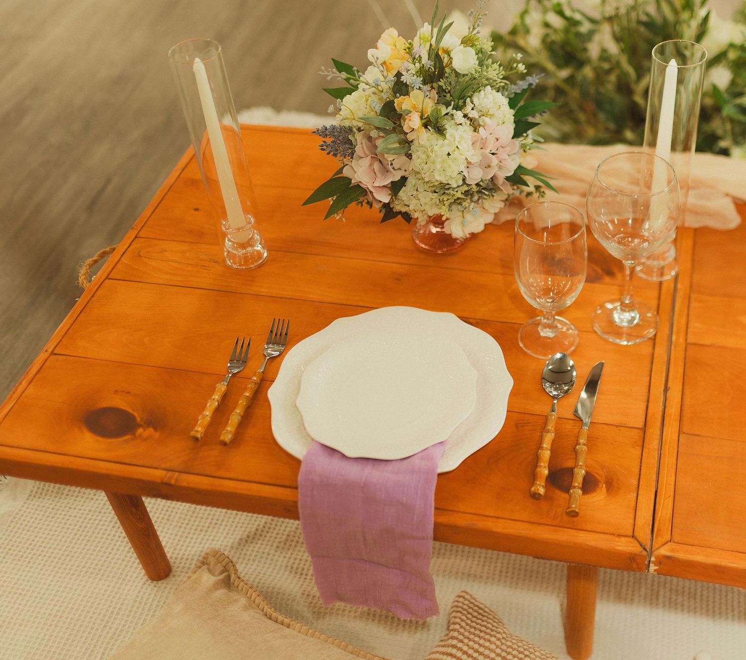 A romantic table setting with a pink napkin, white plate, and floral centerpiece on an orange wooden table.