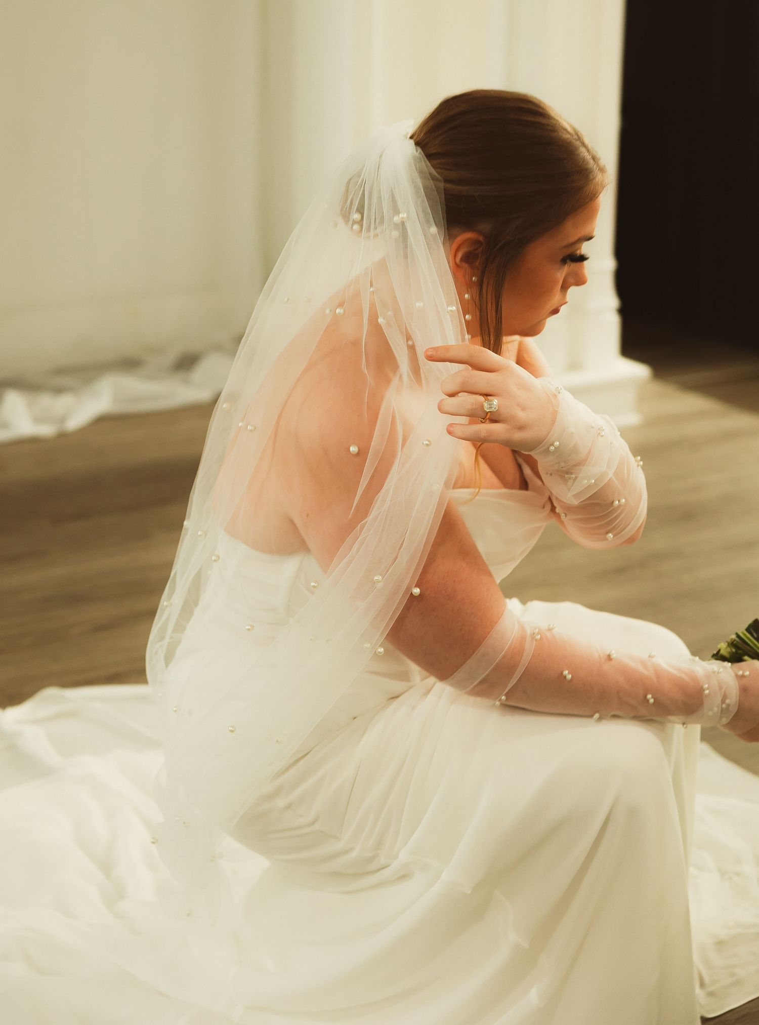 A bride in a flowing white gown and veil looks down pensively in soft window light during a bridal portrait session.