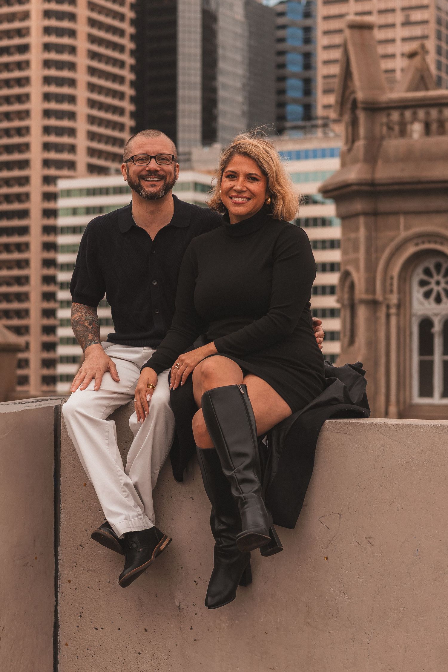 Couple sitting casually on concrete ledge with urban architecture behind them.