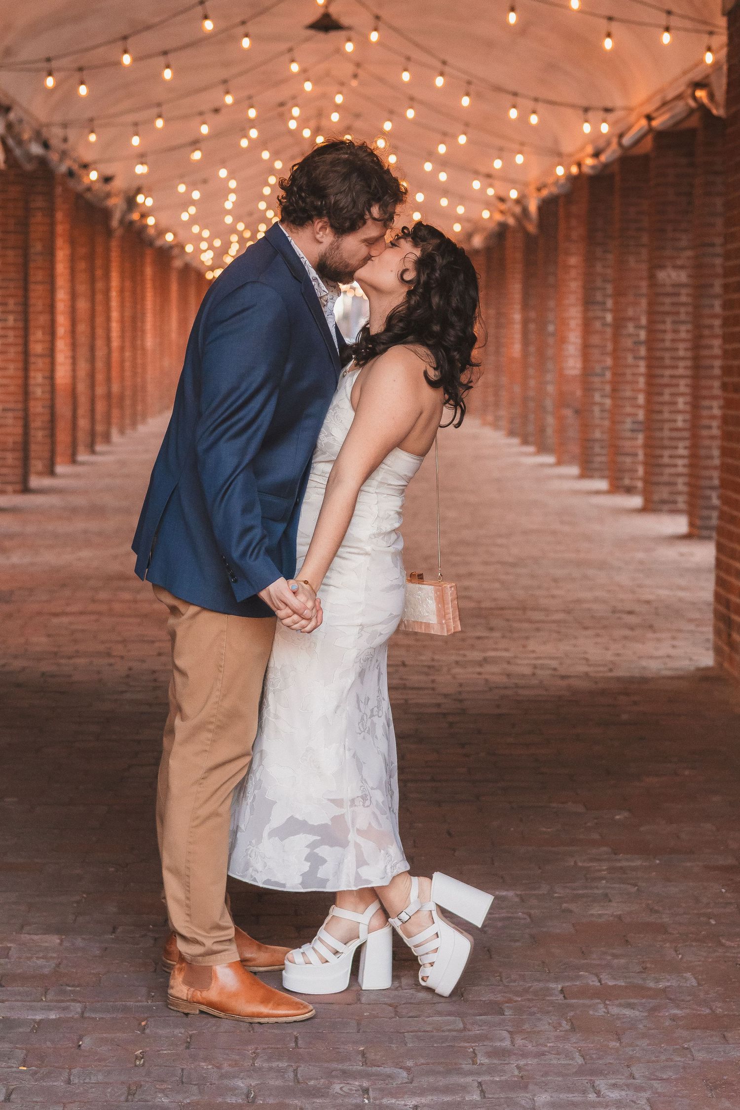 A couple in formal attire sharing a romantic kiss under string lights in a brick corridor.