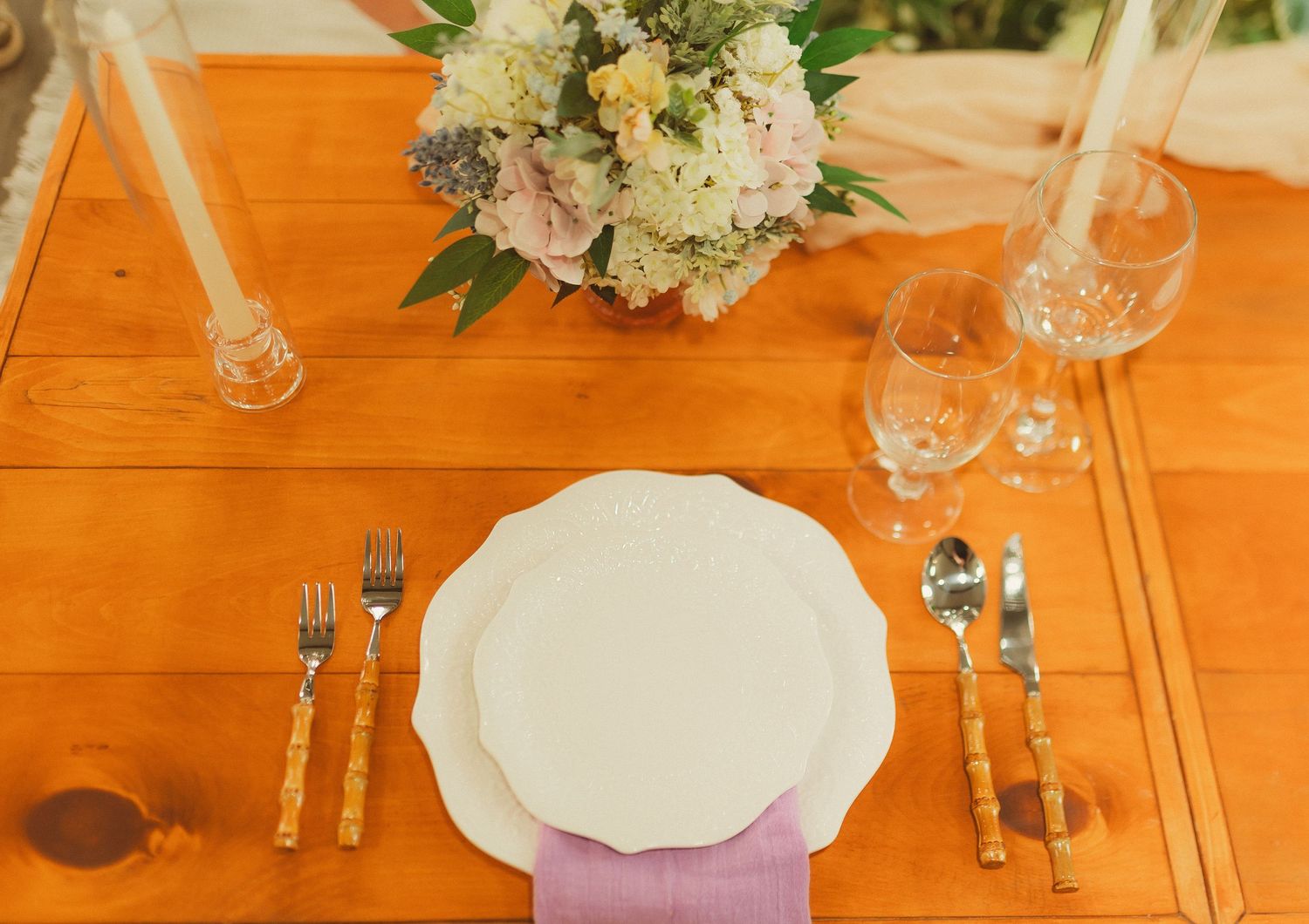 Elegant place setting on wooden table with white plate, silverware, and floral centerpiece.