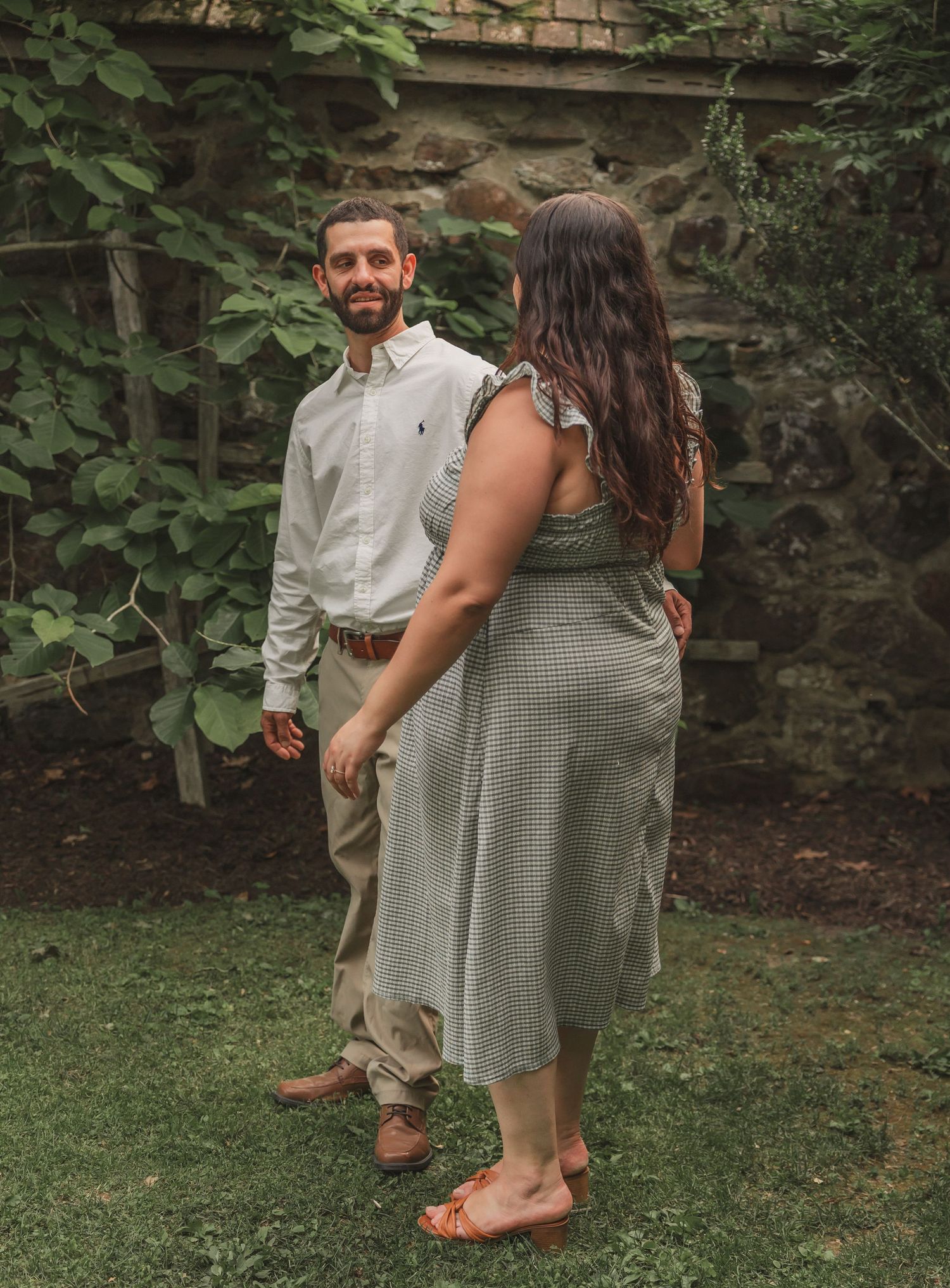 A couple stands together on a lawn surrounded by greenery and a brick wall while sharing a sweet candid moment.