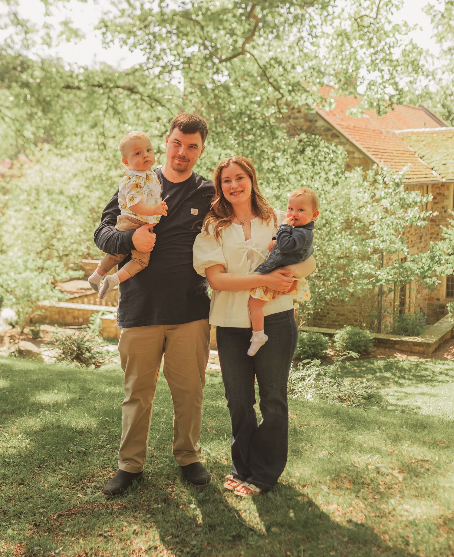 Family stands together in a sun-dappled garden setting with natural bokeh and warm vintage tones.