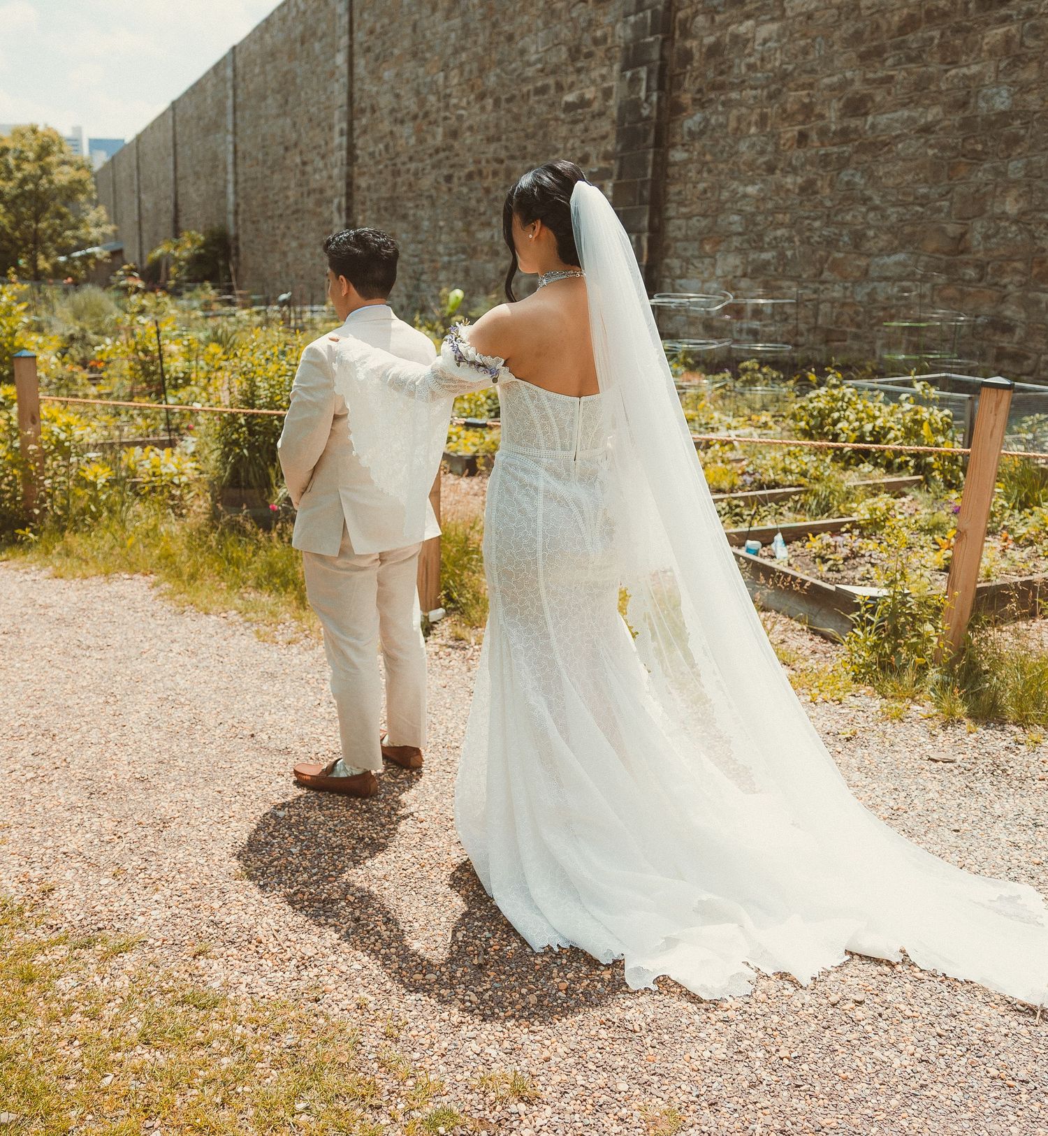 A couple in wedding attire walks along a gravel path with stone wall and garden on a sunny day.