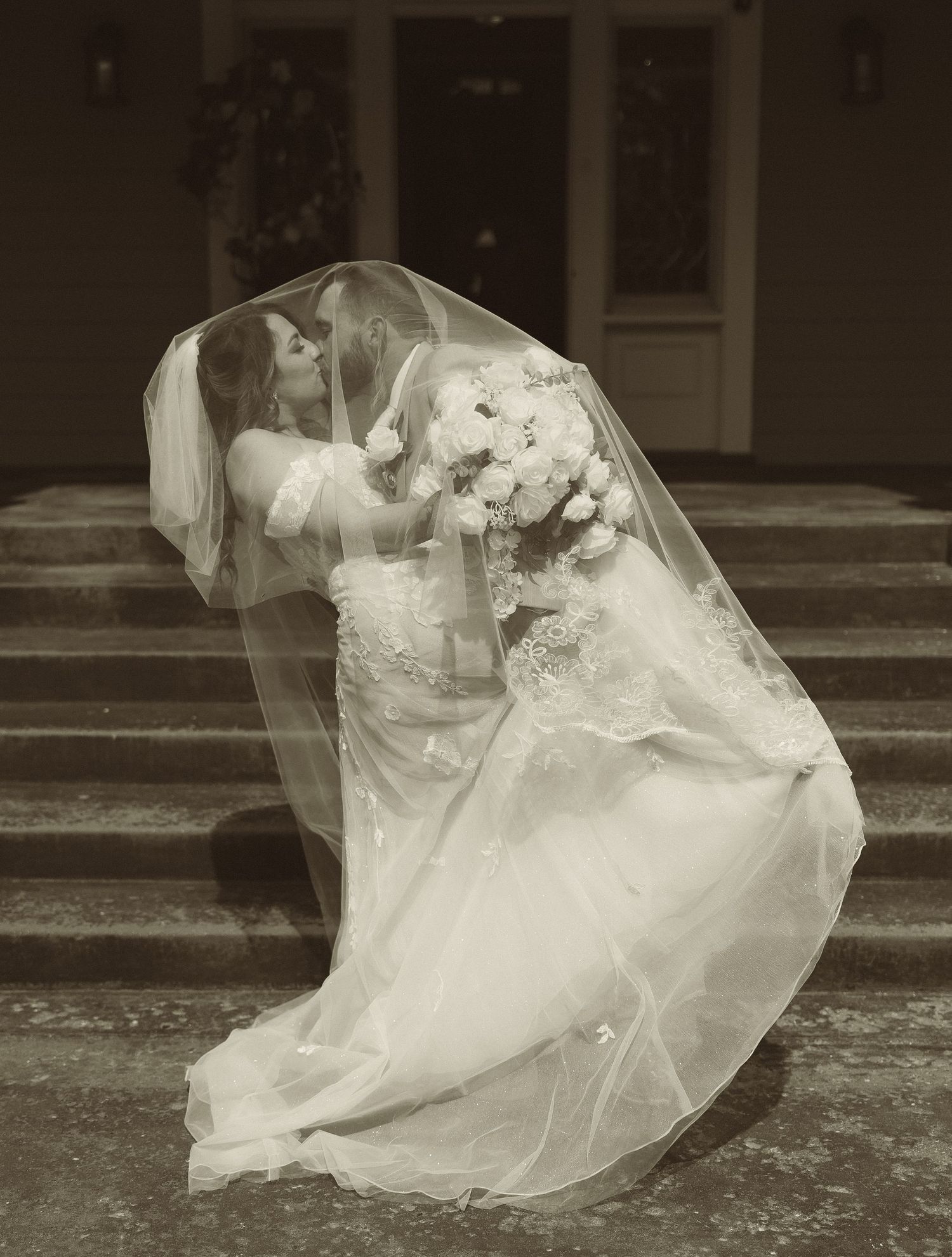 A dramatic black and white wedding photo with flowing veil on stone steps.