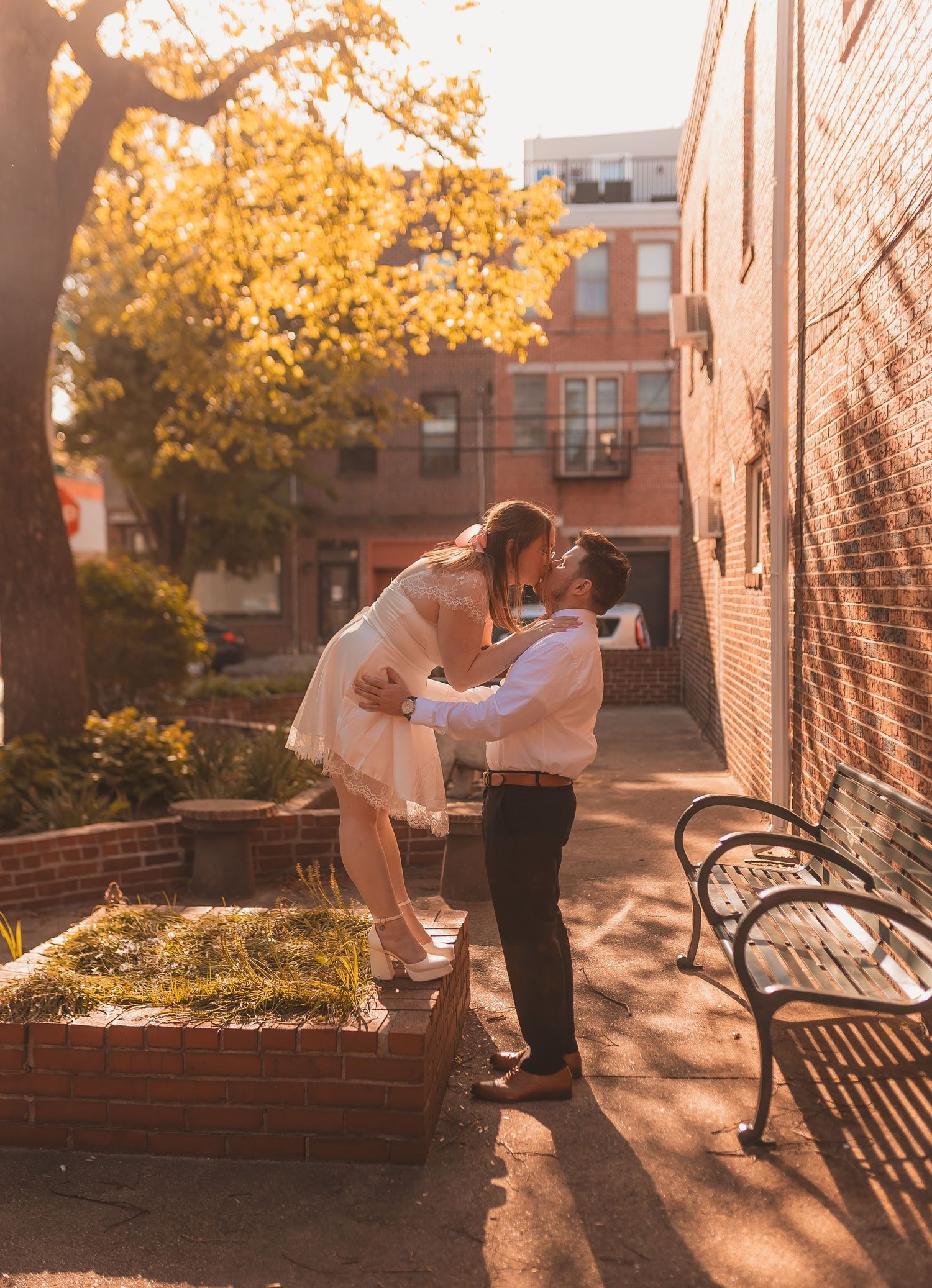 A romantic couple shares an intimate moment near a brick building with autumn foliage.