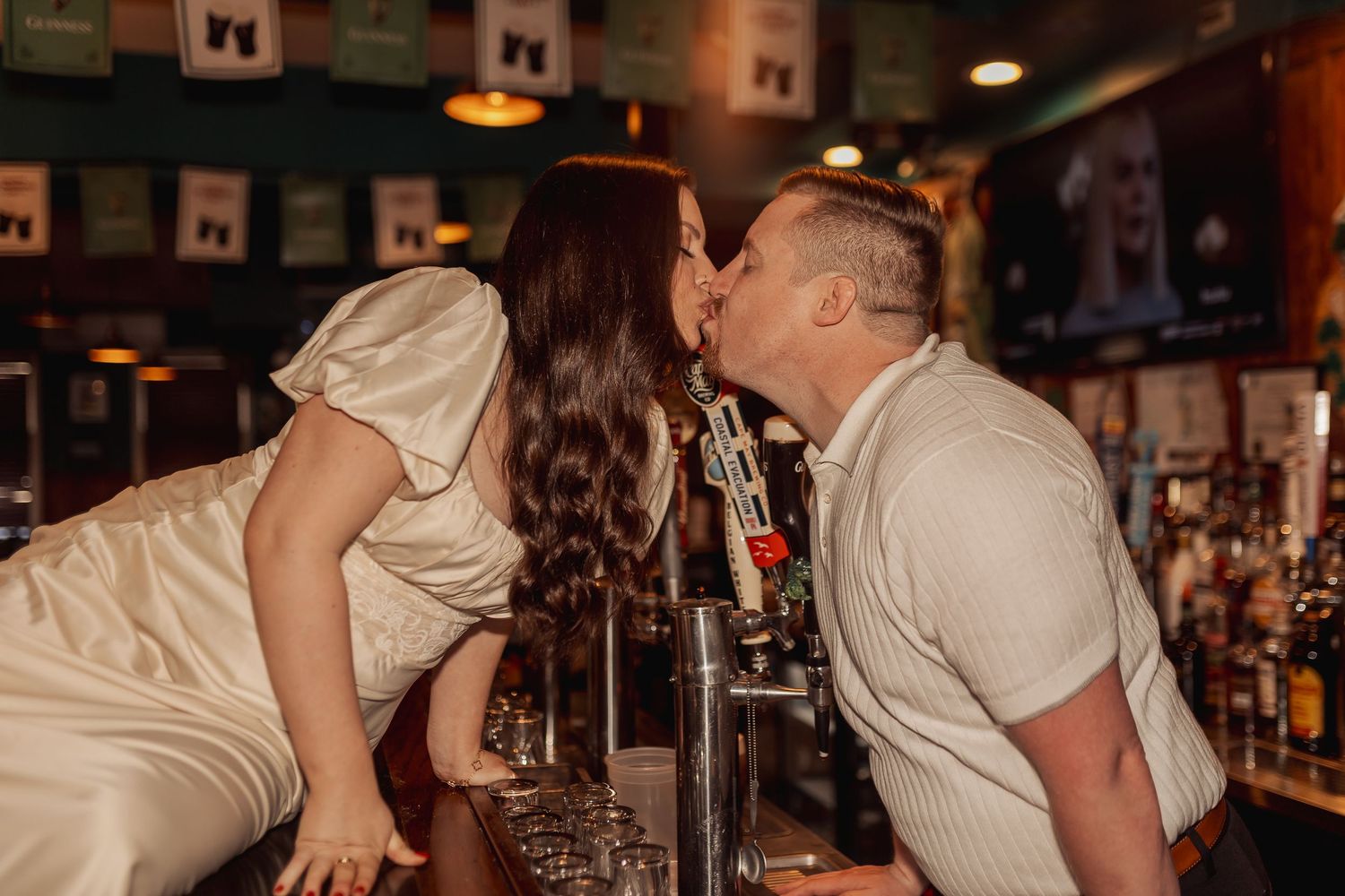 A couple shares an intimate kiss while leaning over a bar counter in a dimly lit pub atmosphere.