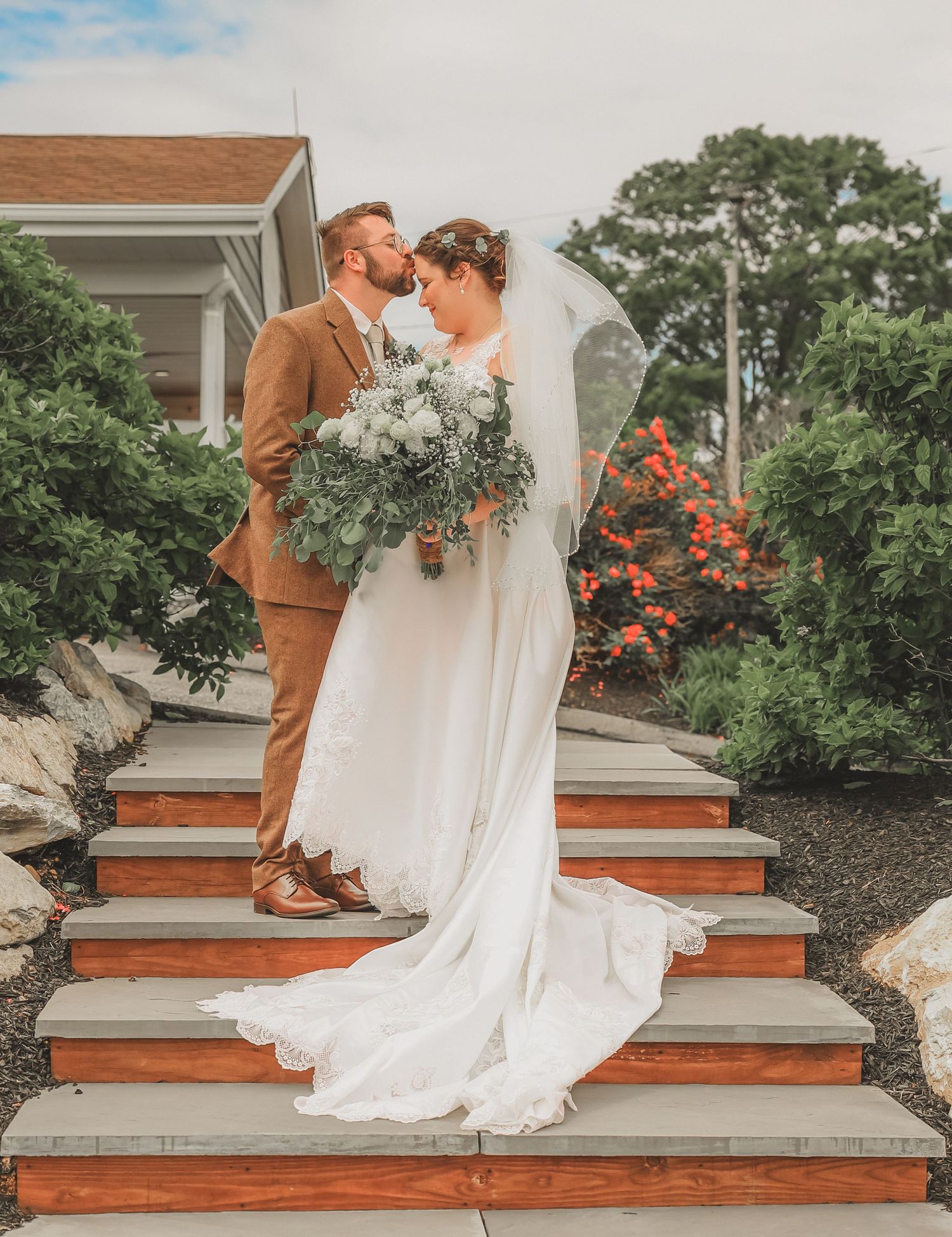 A couple in wedding attire share an intimate moment on wooden steps outside, with a lush garden backdrop.