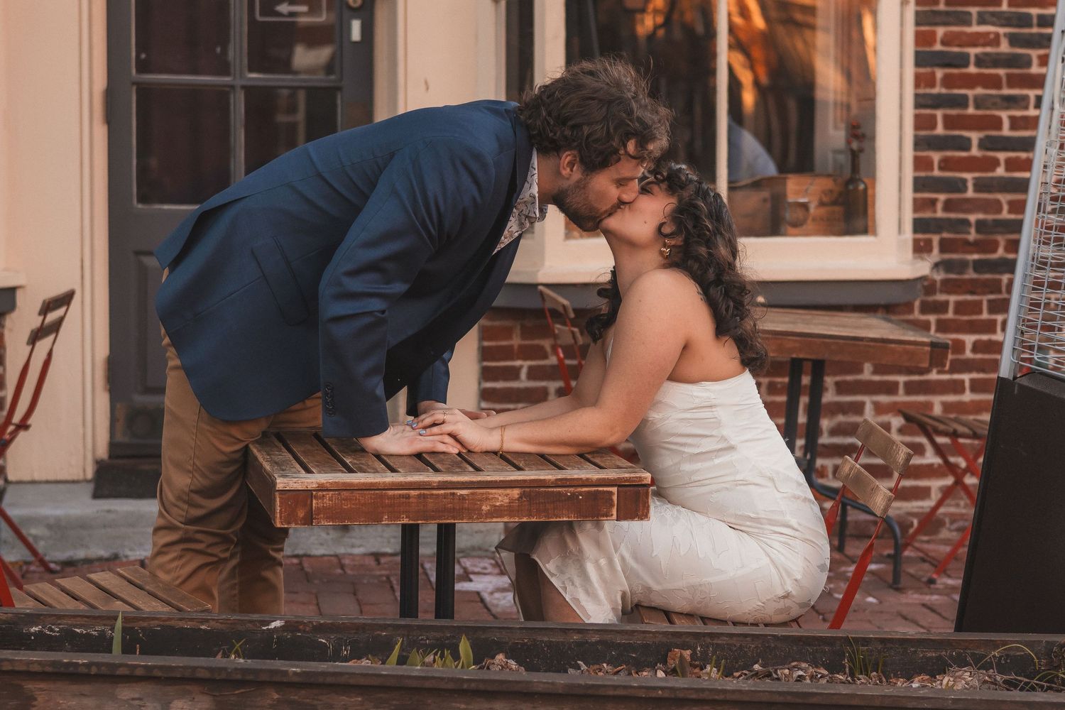 A couple shares a tender moment on a wooden bench outside a brick building.