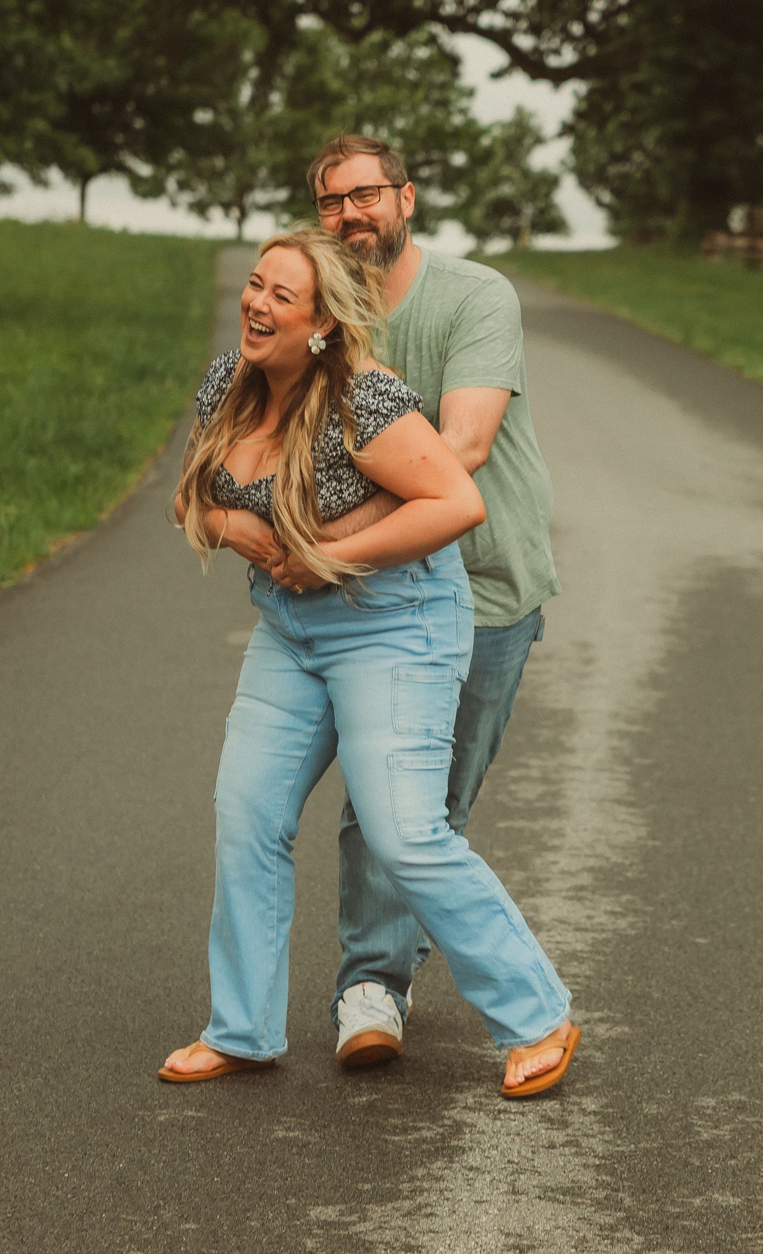 A playful couple dancing together on a paved pathway surrounded by green grass.