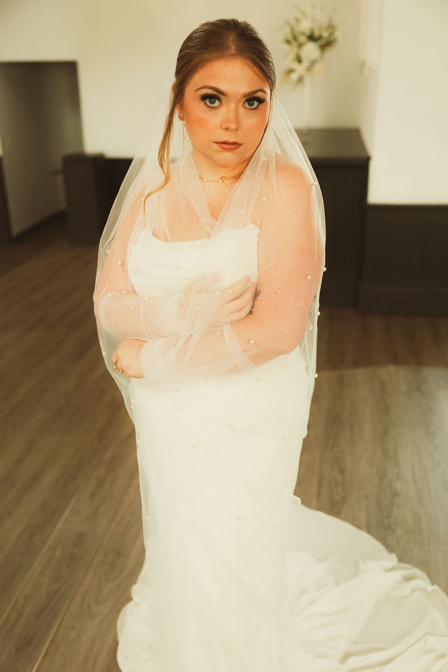 A bride in an elegant white mermaid-style wedding gown and delicate veil poses against a hardwood floor backdrop.