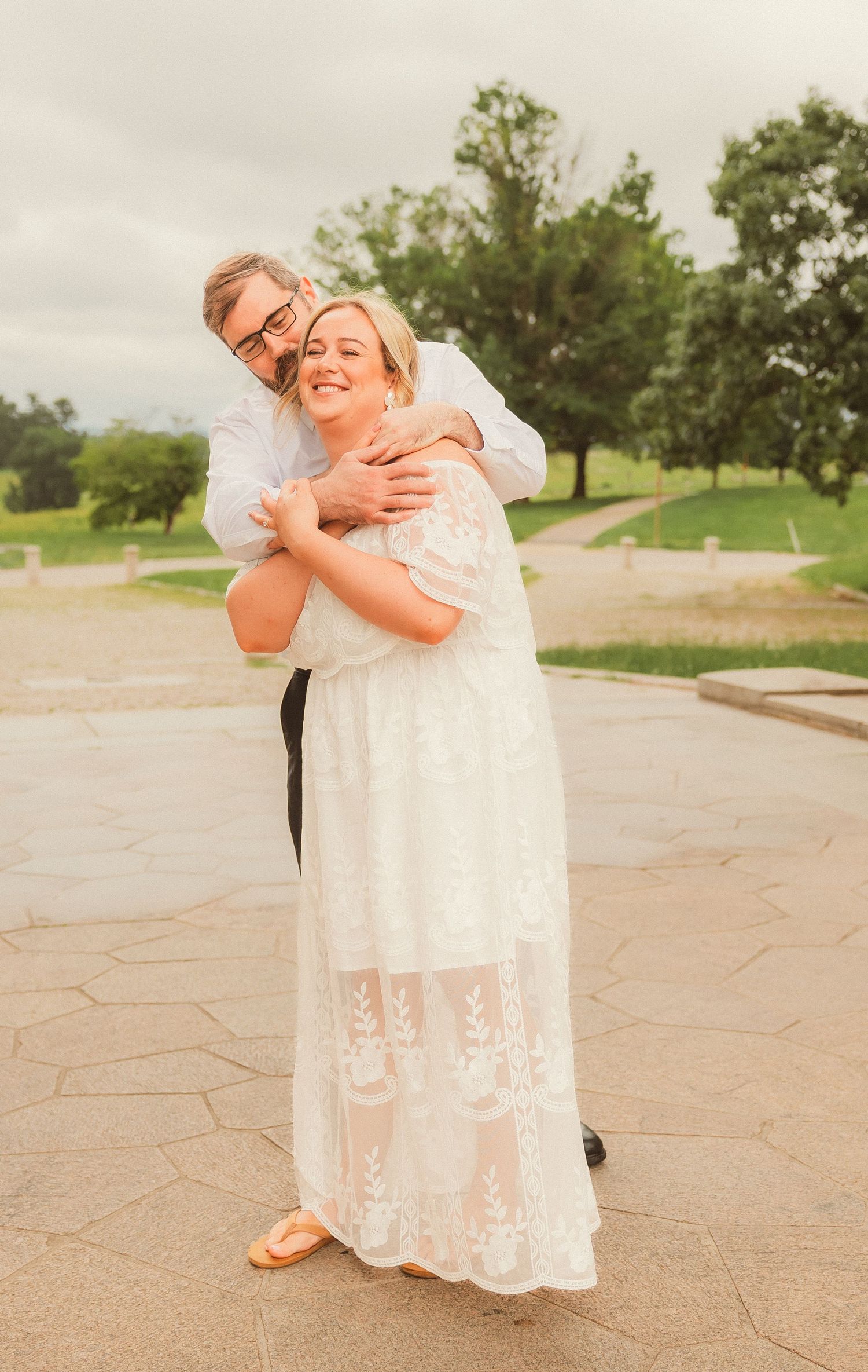 A couple shares a joyful embrace on a stone walkway surrounded by green trees on an overcast day.