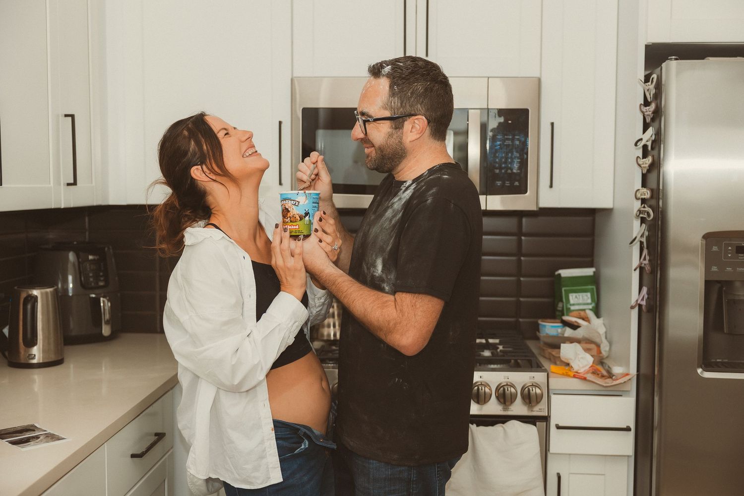 Two people share an intimate moment while feeding each other snacks in a bright modern kitchen.