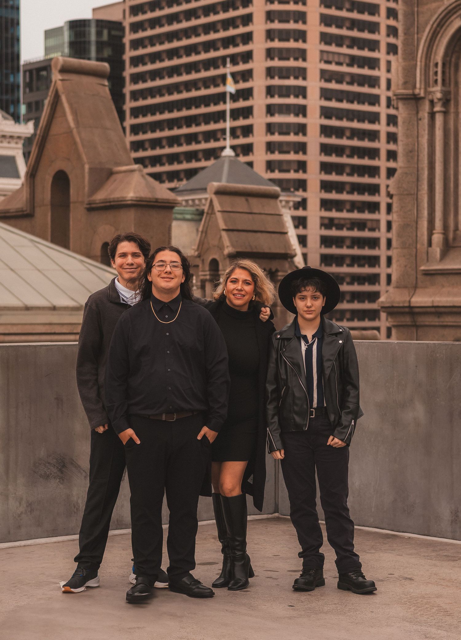 Four friends in dark clothing pose against an urban rooftop backdrop with historic architecture and modern buildings.
