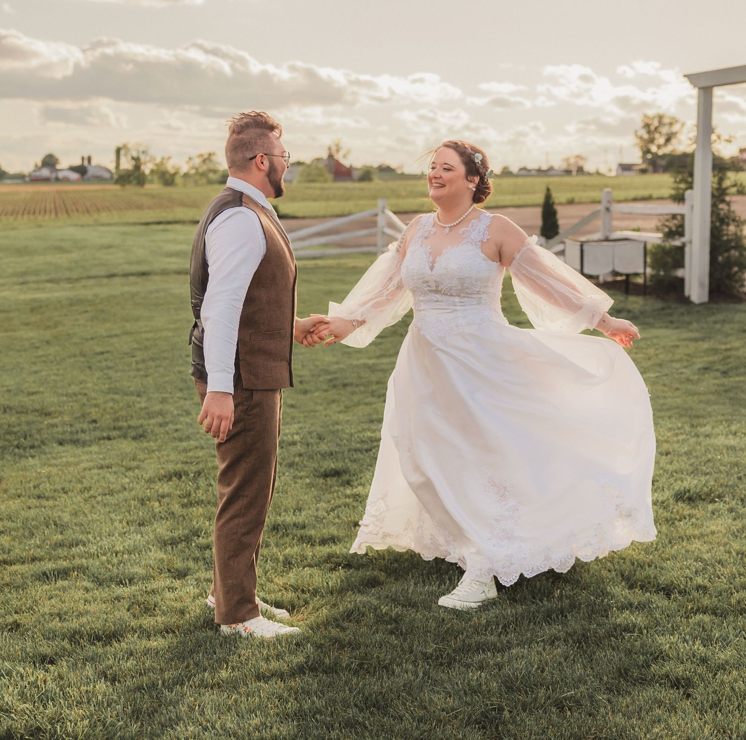 A bride in a flowing white dress twirls joyfully on a grassy field at sunset while her groom watches adoringly.