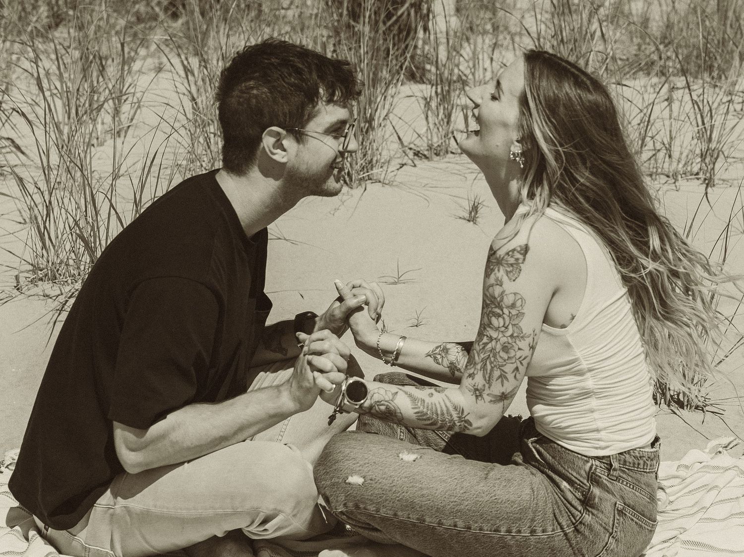 A couple sits on a sandy beach together sharing a tender moment in black and white.