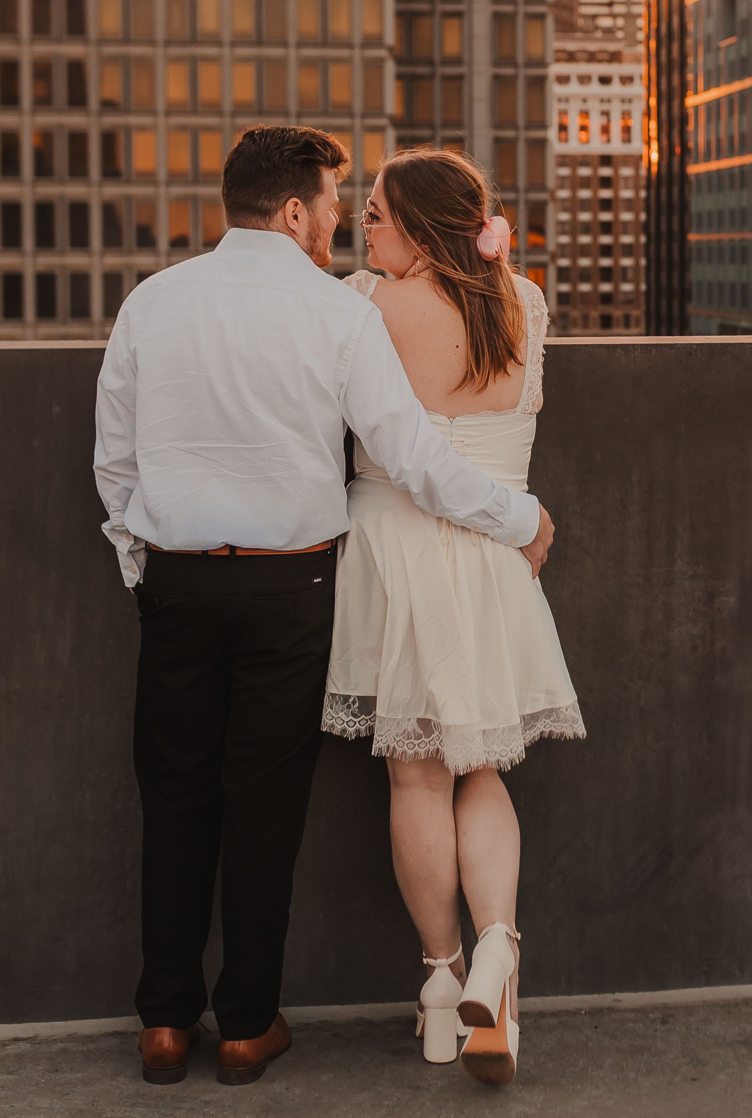 A couple in formal attire stands together against a concrete wall overlooking the city at dusk.