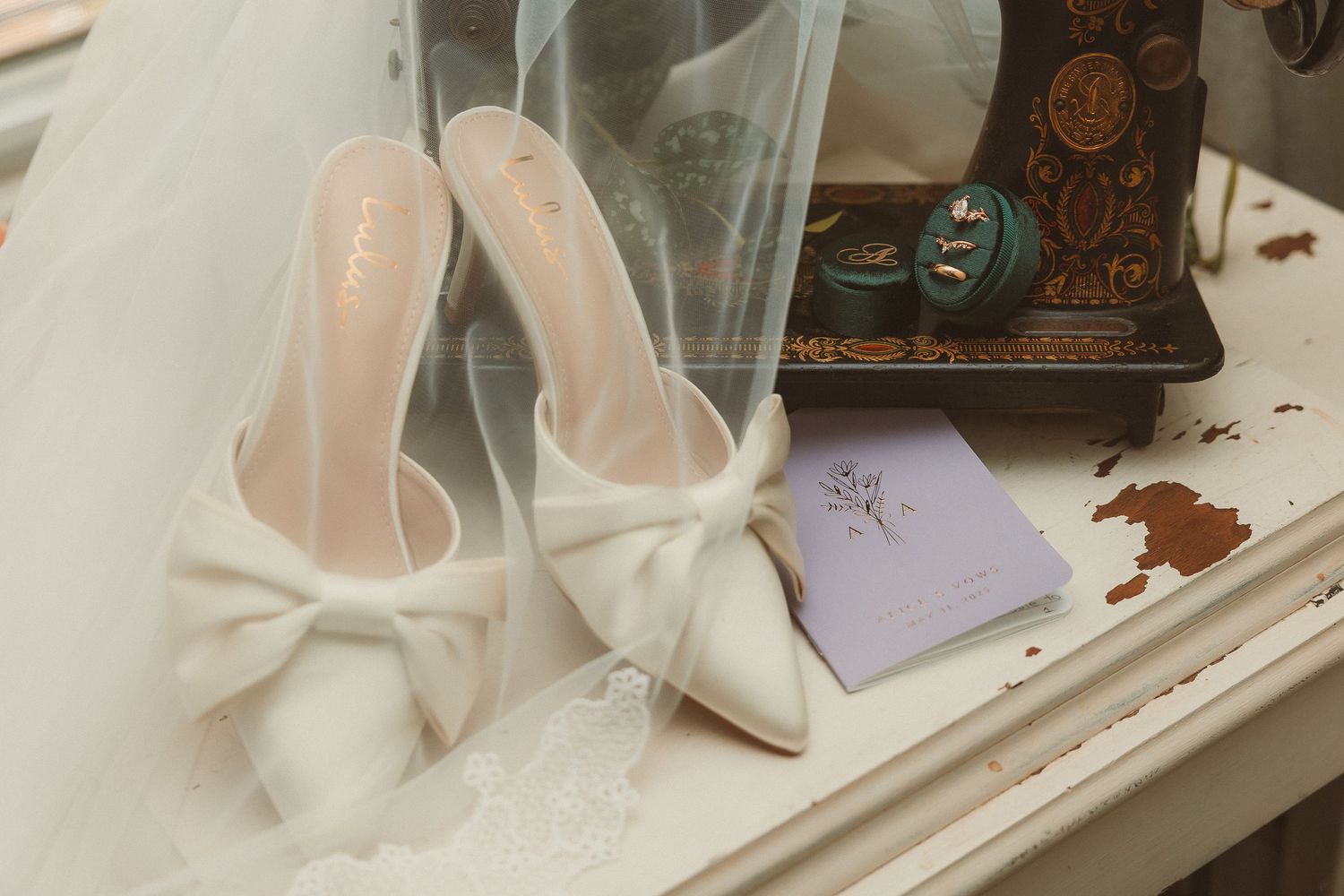 Elegant white bow-adorned bridal shoes rest on a vintage vanity beside a lilac invitation card and antique sewing machine.