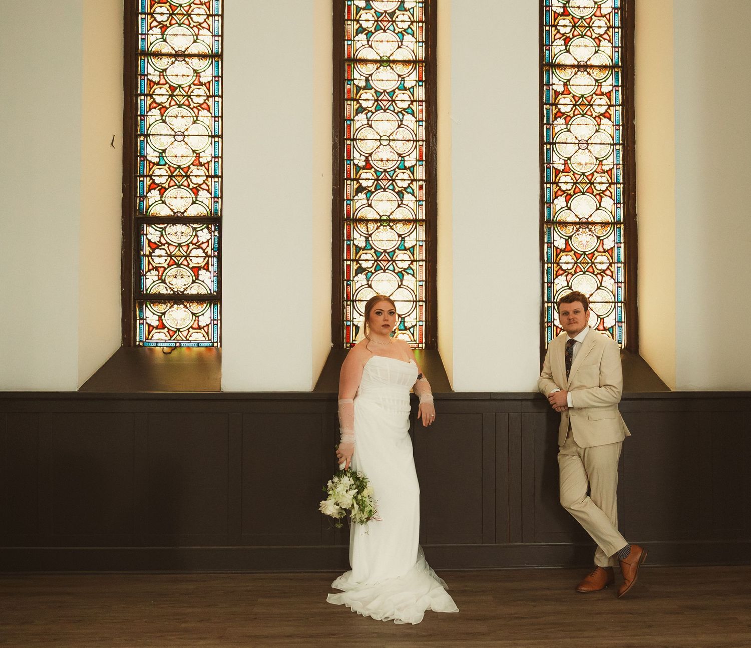 A wedding scene in a church with a bride and groom walking past stained glass windows in warm lighting.