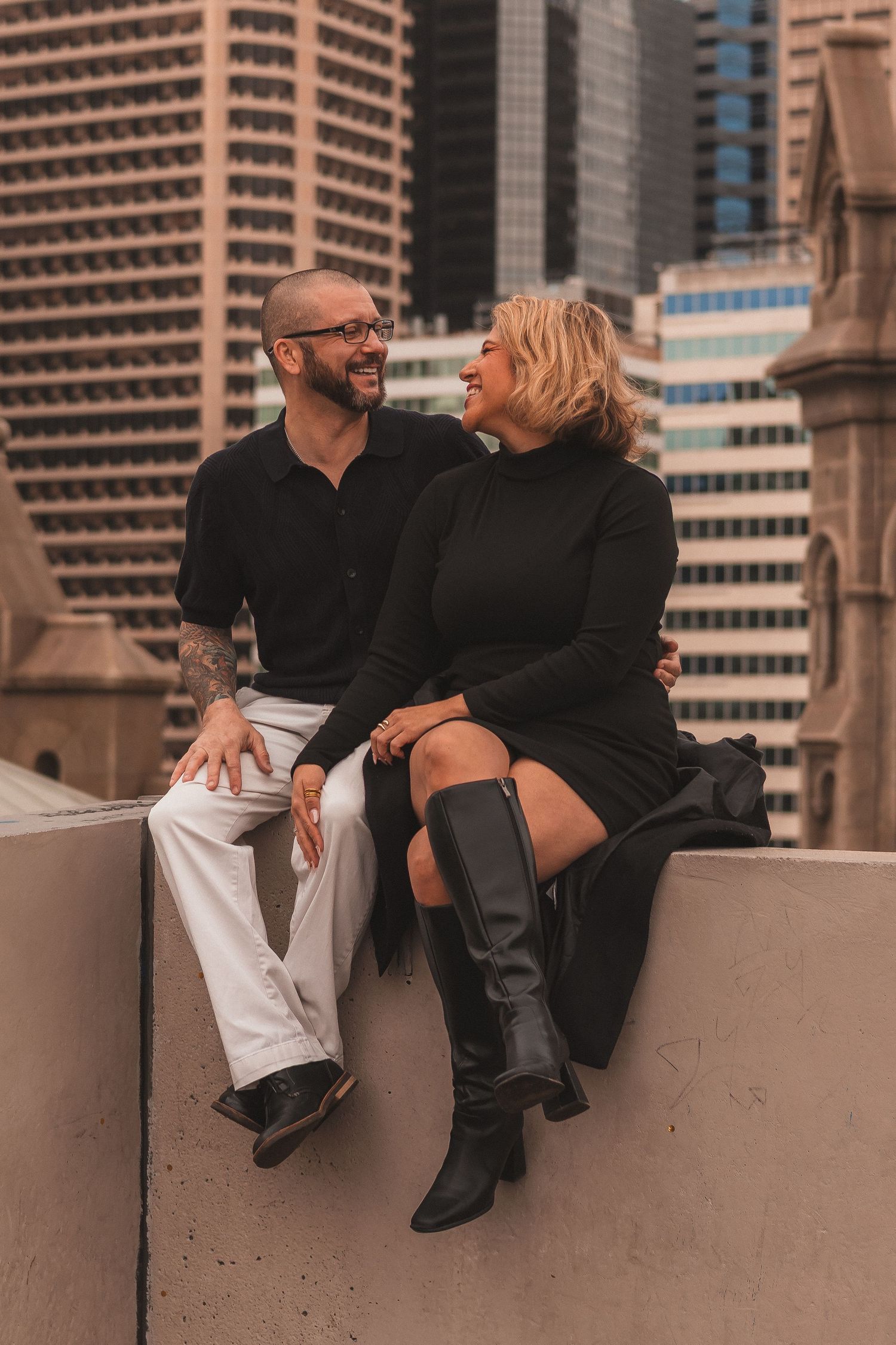 Couple sharing tender moment while sitting on concrete wall near downtown buildings.