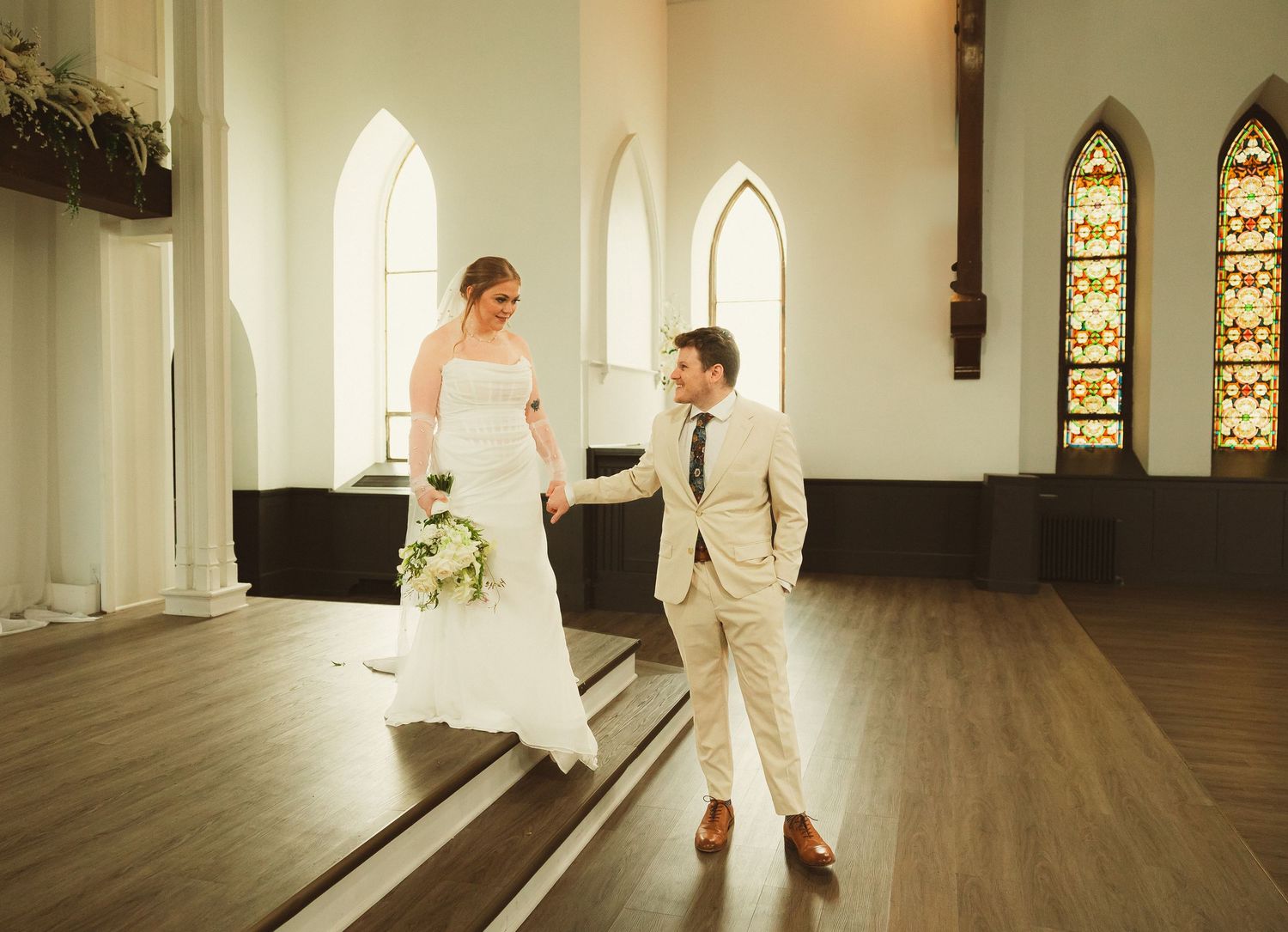 A couple shares a joyful moment at their wedding ceremony in a bright, historic chapel with arched stained glass windows.